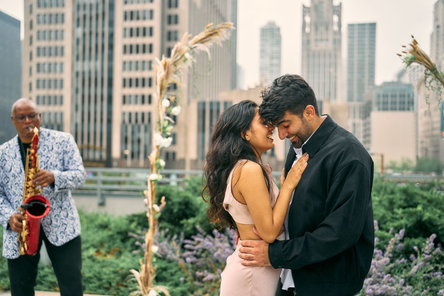 A romantic couple embraces in an urban garden while a saxophone player performs in the background with city skyline views.
