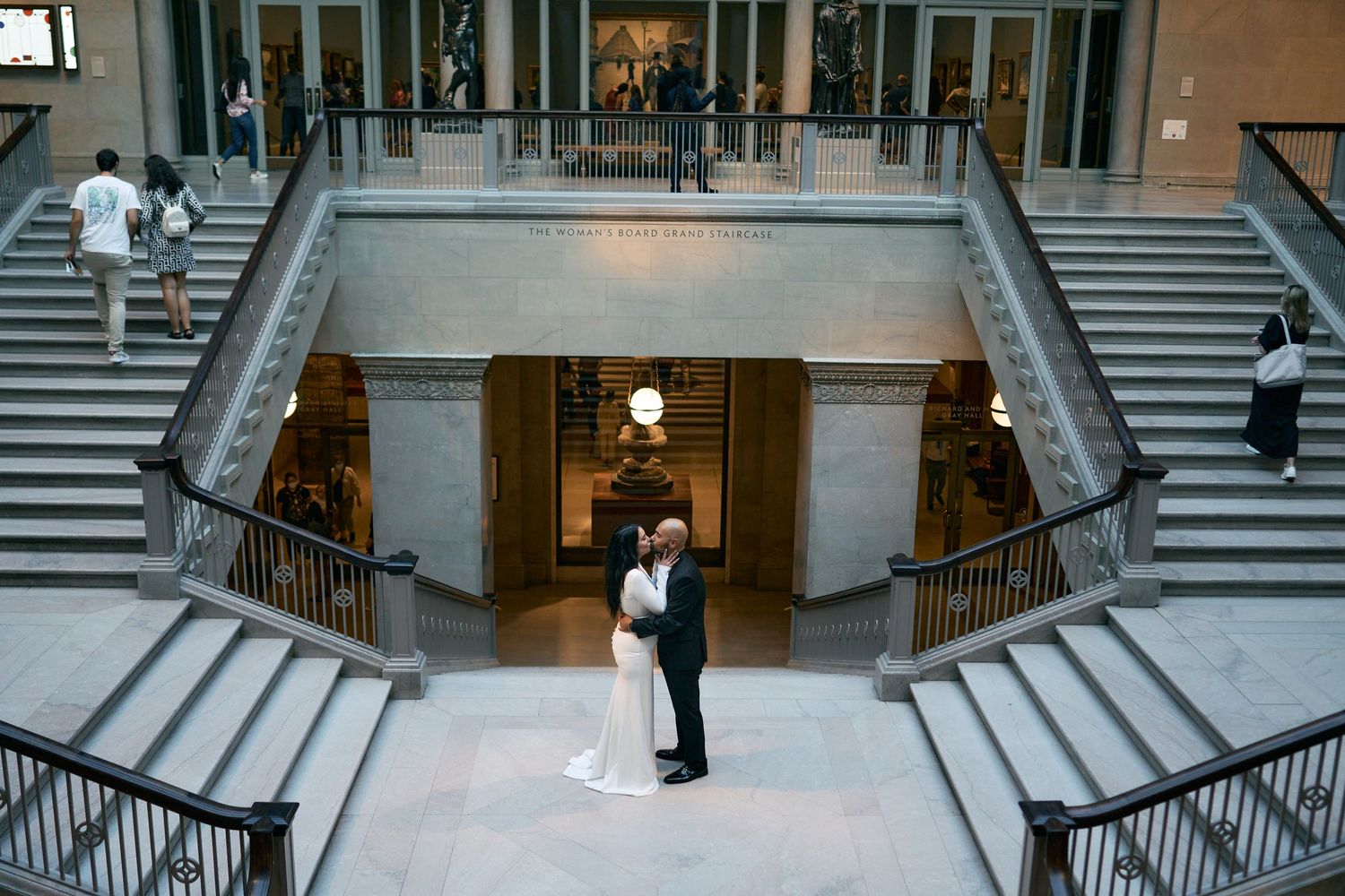 Two people share an intimate embrace on grand stone staircase in elegant indoor setting with dramatic lighting.