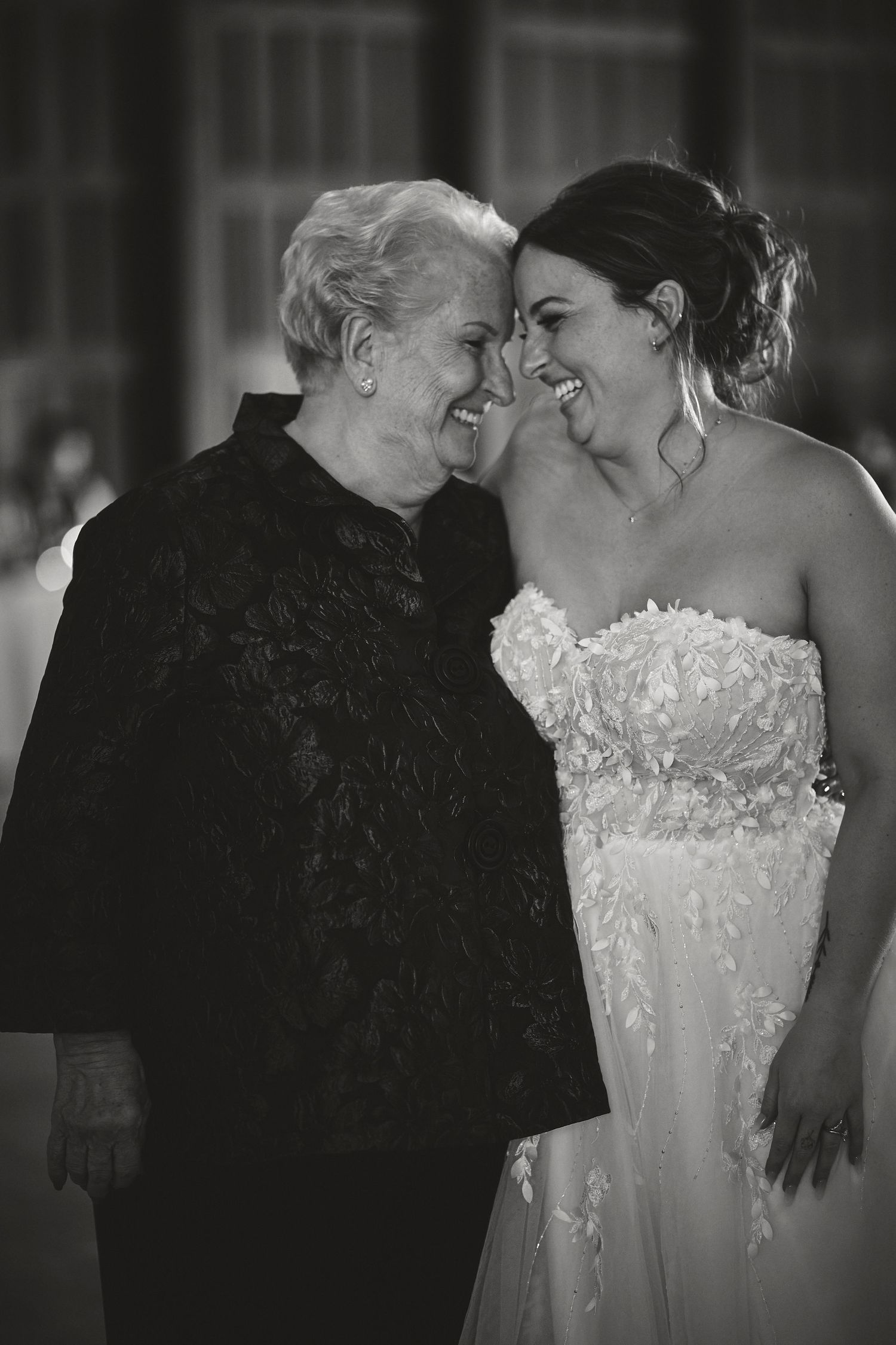 A black and white photo shows an intimate moment between a bride in a strapless gown and an elderly woman in dark clothing.