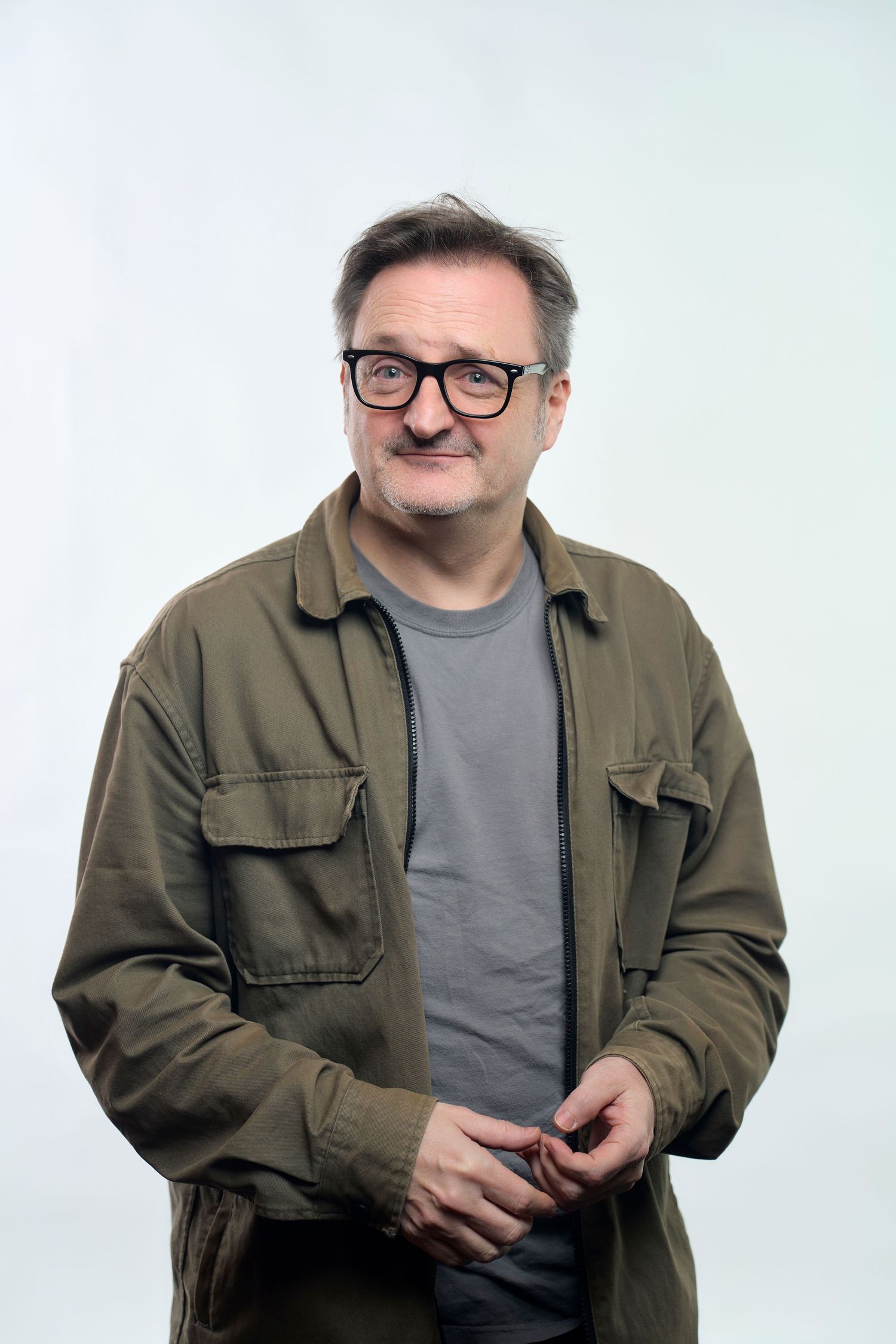 Portrait of a smiling person wearing glasses and an olive green utility jacket against a light background.