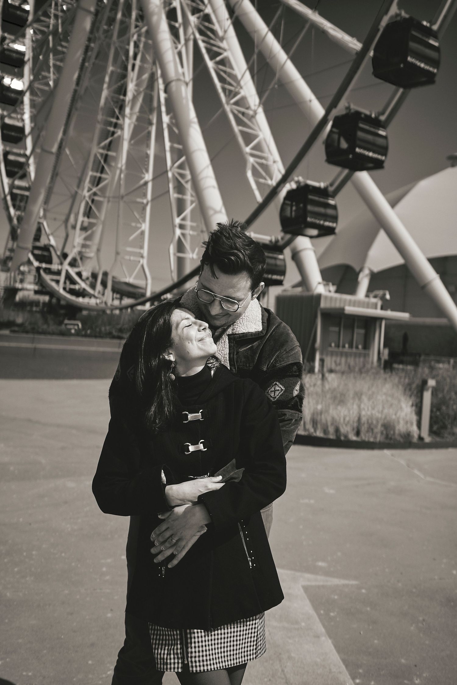 A couple sharing a romantic moment in front of a large ferris wheel in black and white photography.