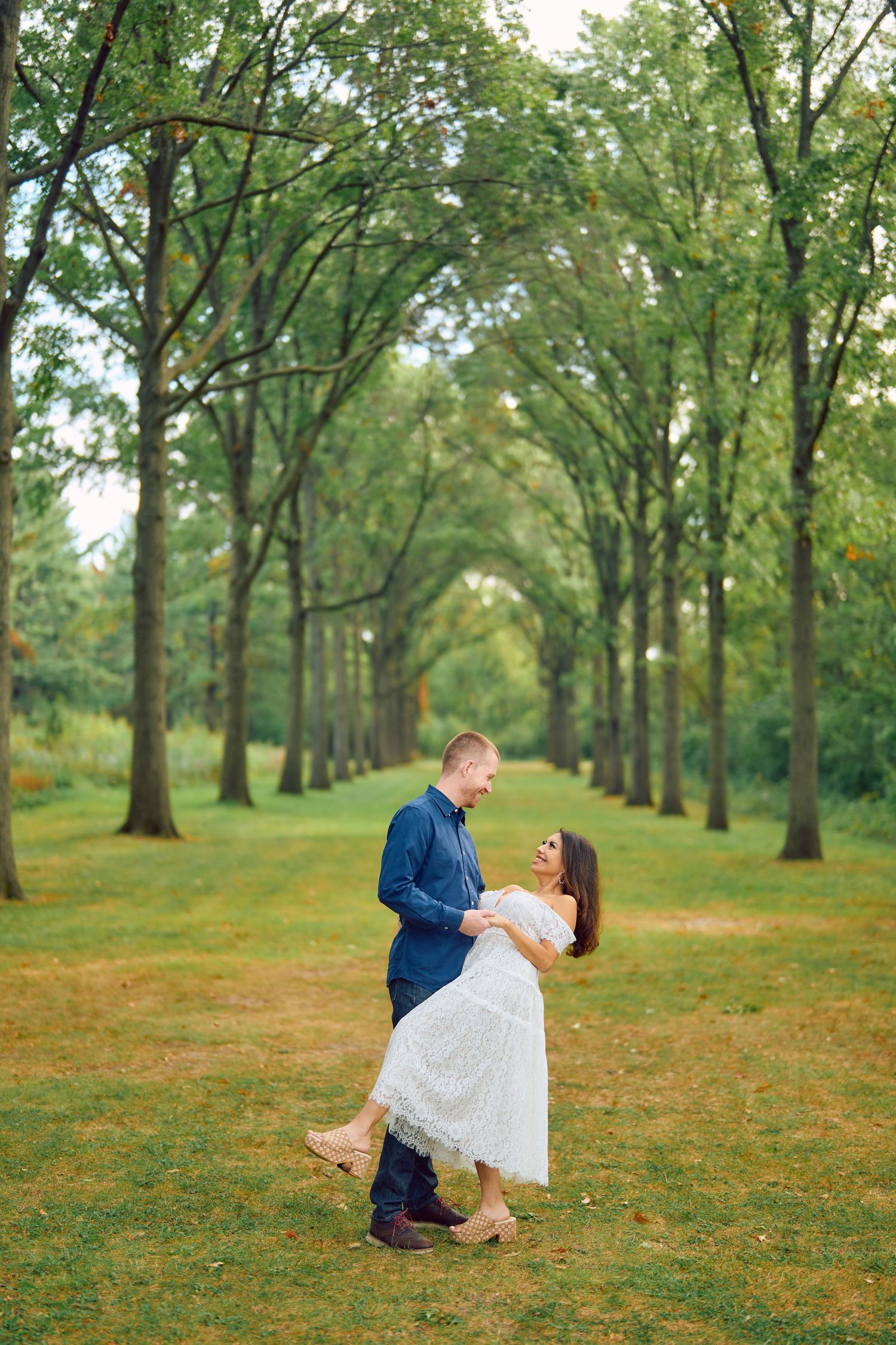 A couple shares a romantic moment under a canopy of tall trees creating a natural green tunnel on a grassy path.