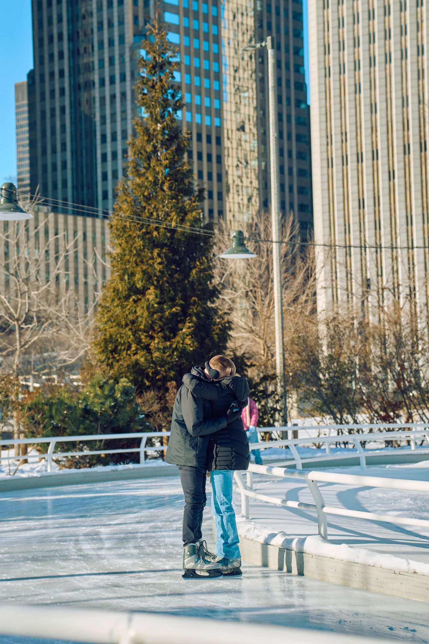 Two people ice skating together at an urban rink with skyscrapers in the background on a sunny winter day.