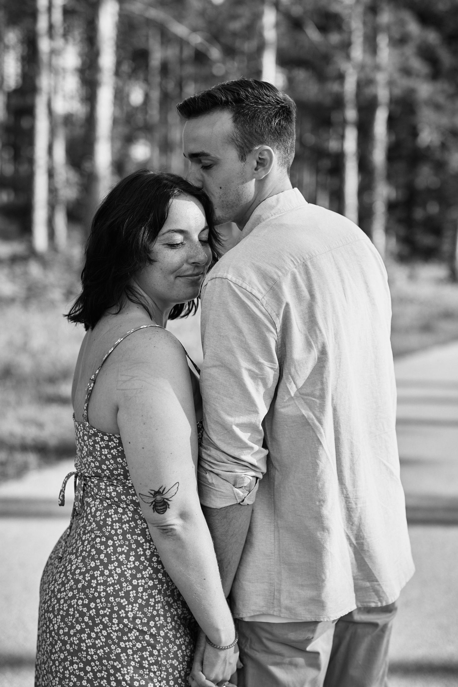 A romantic black and white portrait of a couple embracing outdoors in a forest setting, she wearing a floral dress.