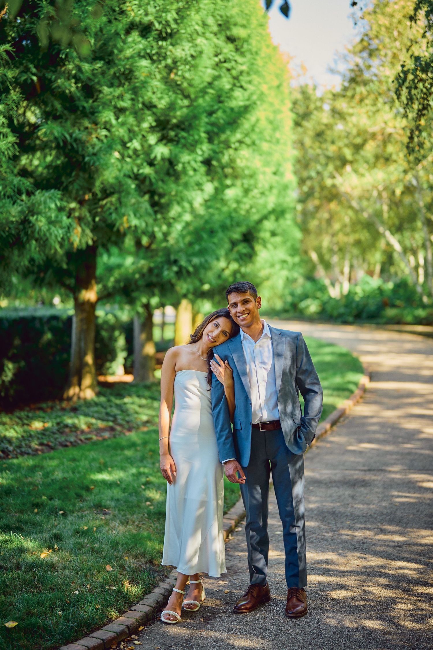 A couple standing together on a tree-lined path on a sunny day, she in a white dress and he in a blue suit.