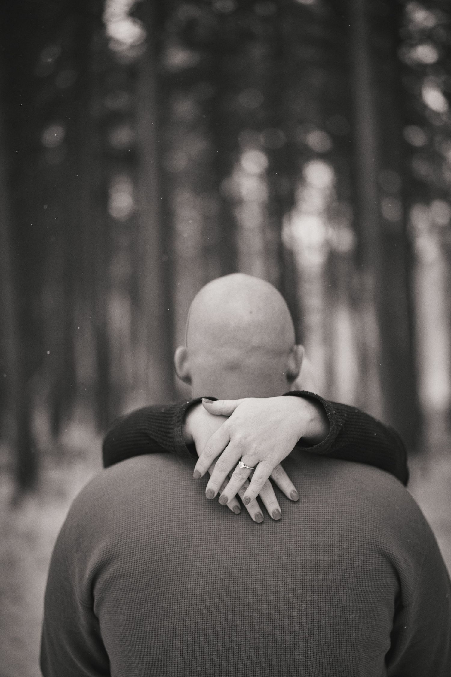Black and white photo of someone being hugged from behind with hands visible on their shoulders in a forest setting.