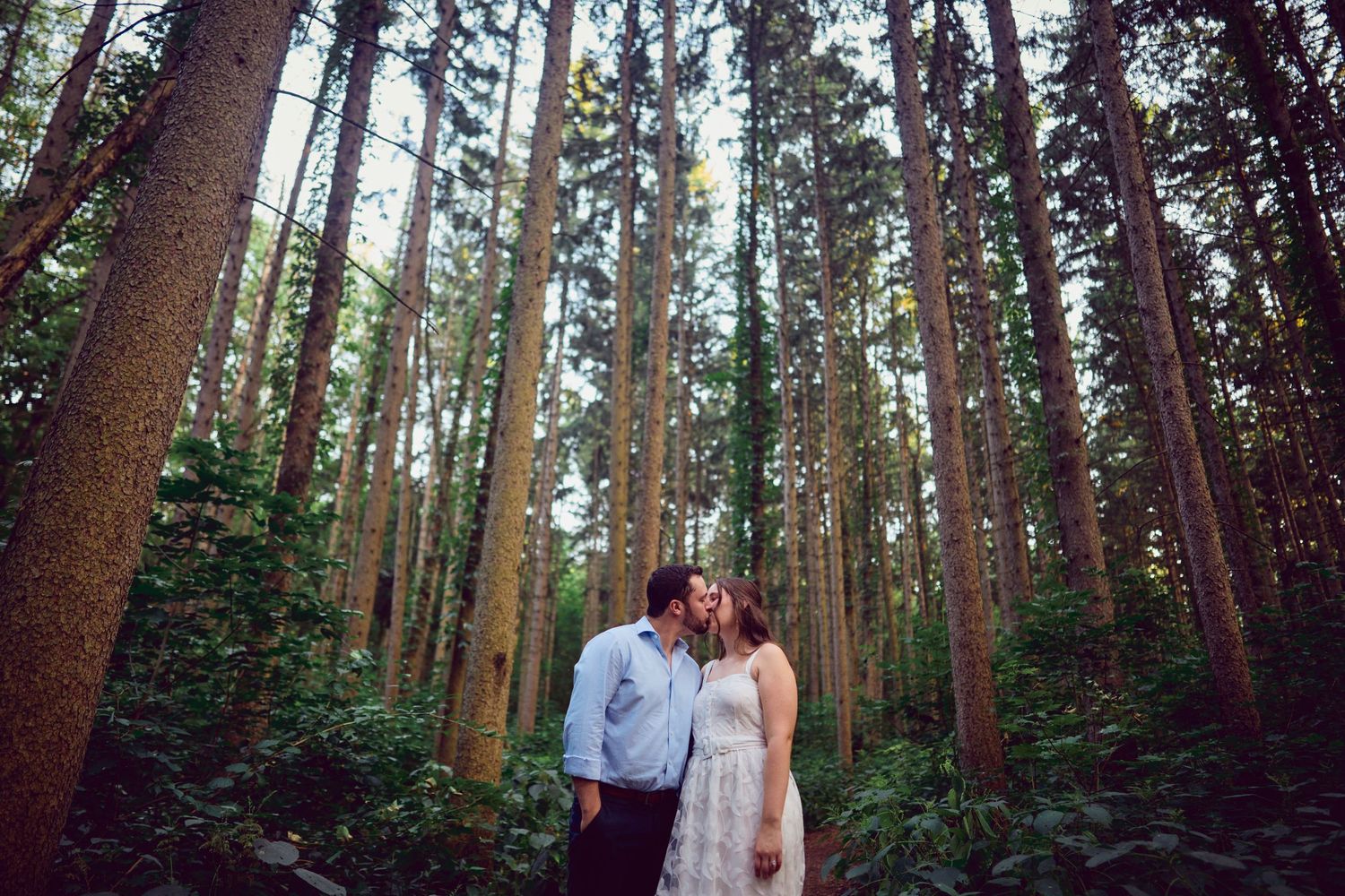 Two people pose together in a romantic forest setting surrounded by tall evergreen trees.