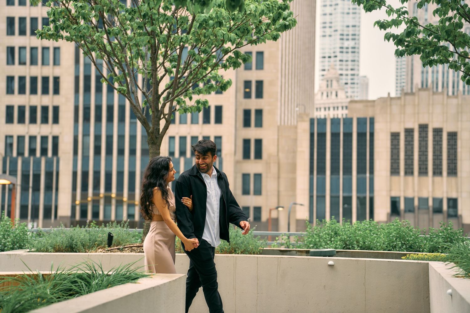 Two people walking and conversing near urban office buildings on a rooftop garden area in a city setting.