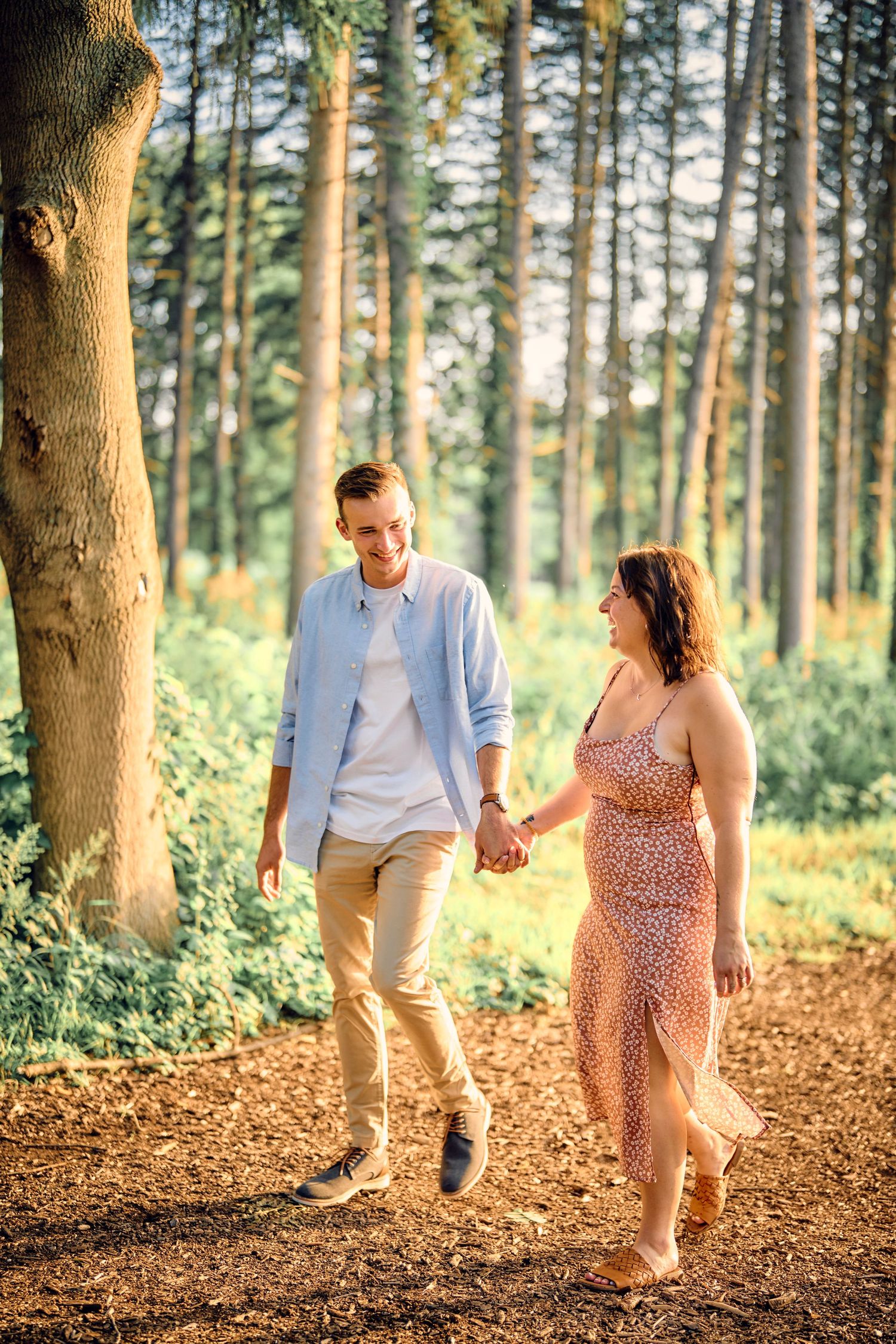 A couple walks together on a sunlit forest path while holding hands in a romantic woodland setting.