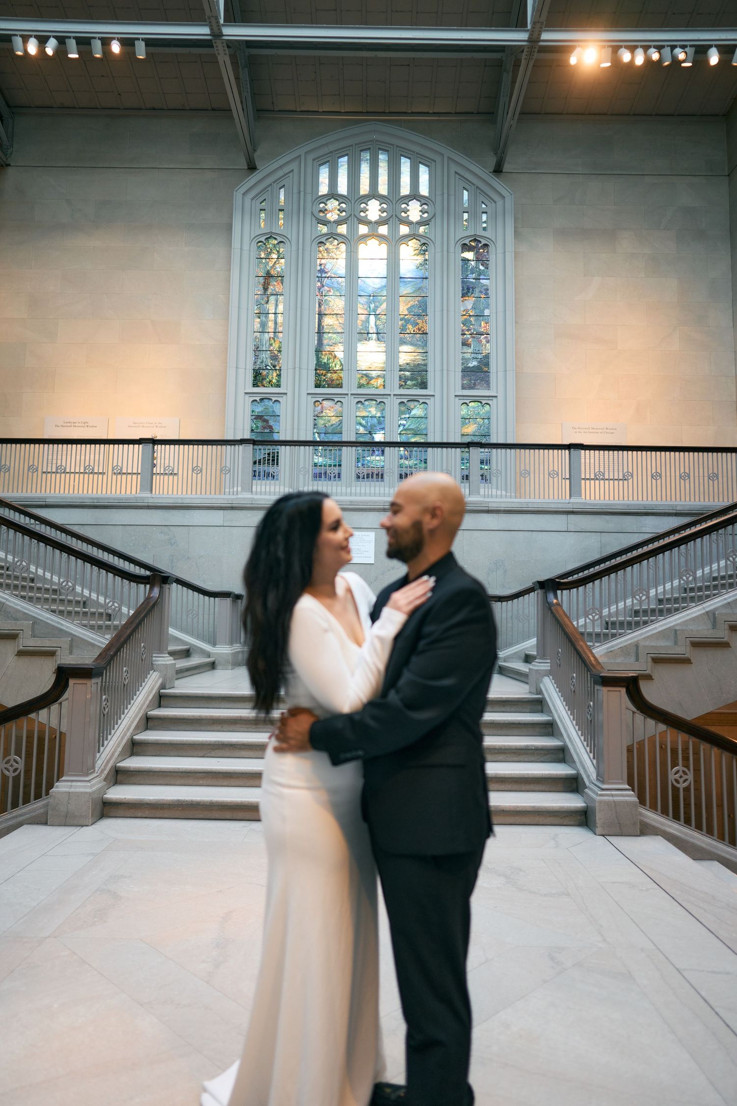 A romantic wedding couple embraces on an elegant staircase beneath a grand stained glass window.