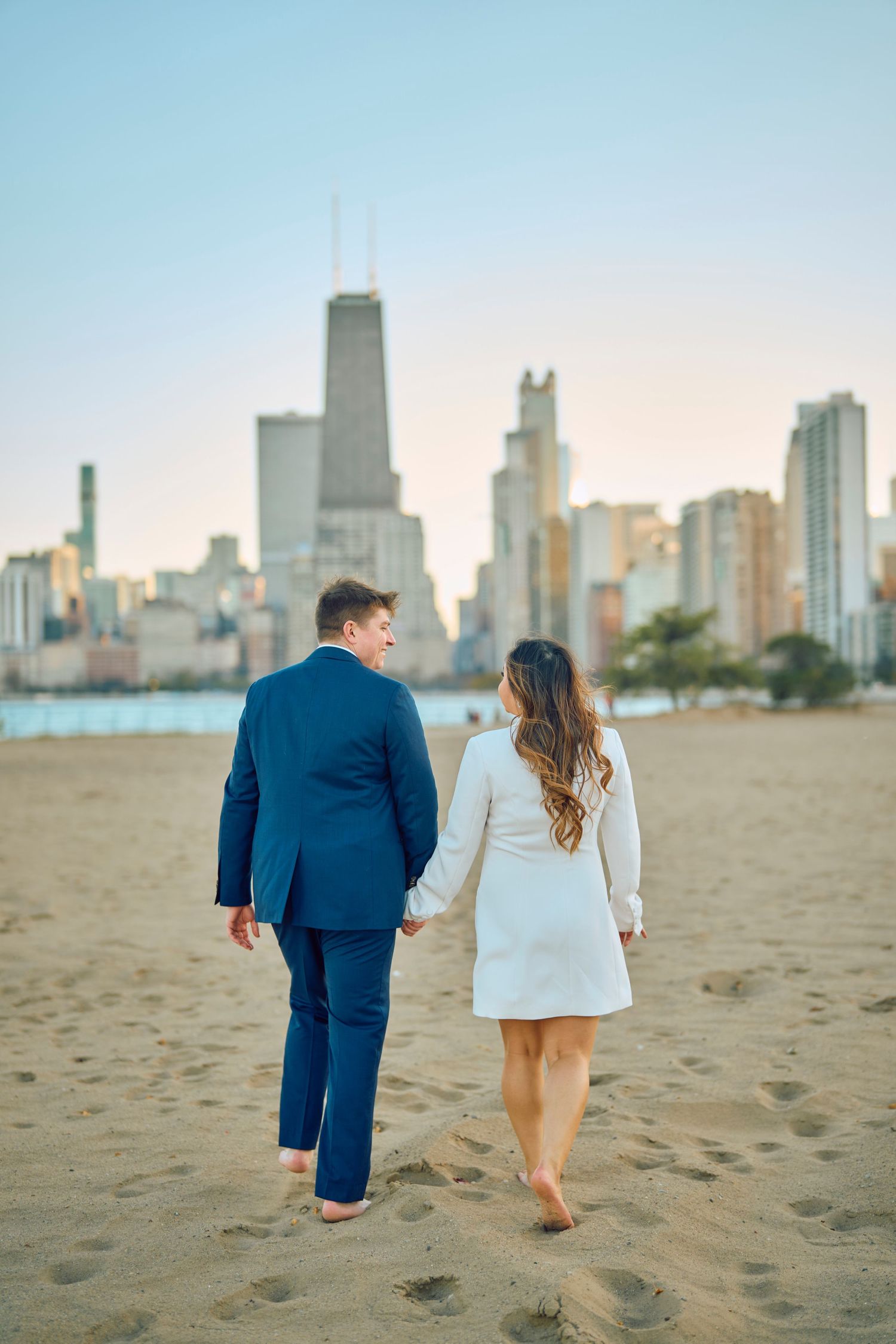 Couple walking barefoot on North Avenue Beach with Chicago skyline and John Hancock building in background at sunset.