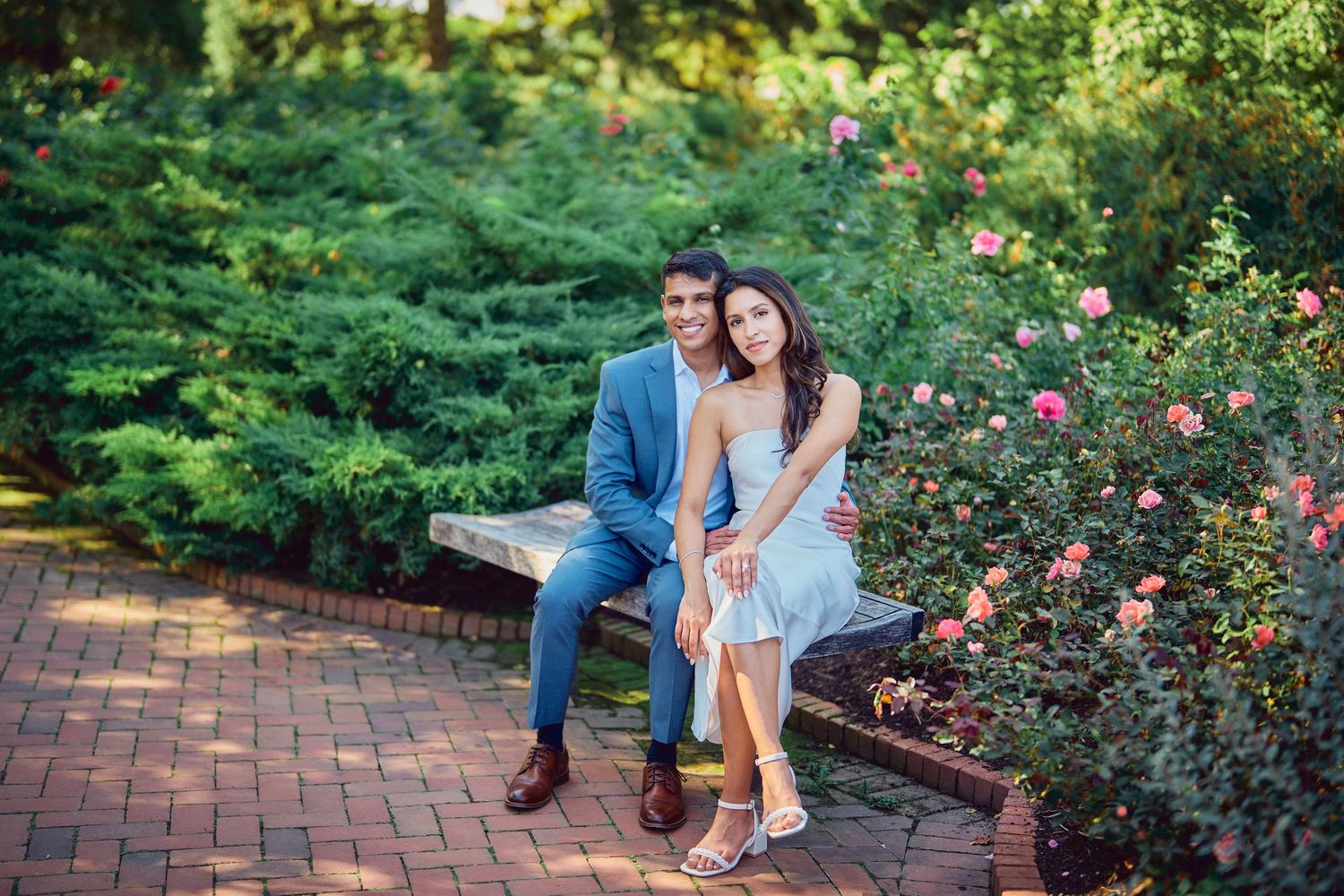 A couple sits together on a brick bench surrounded by lush gardens and blooming roses on a summer day.