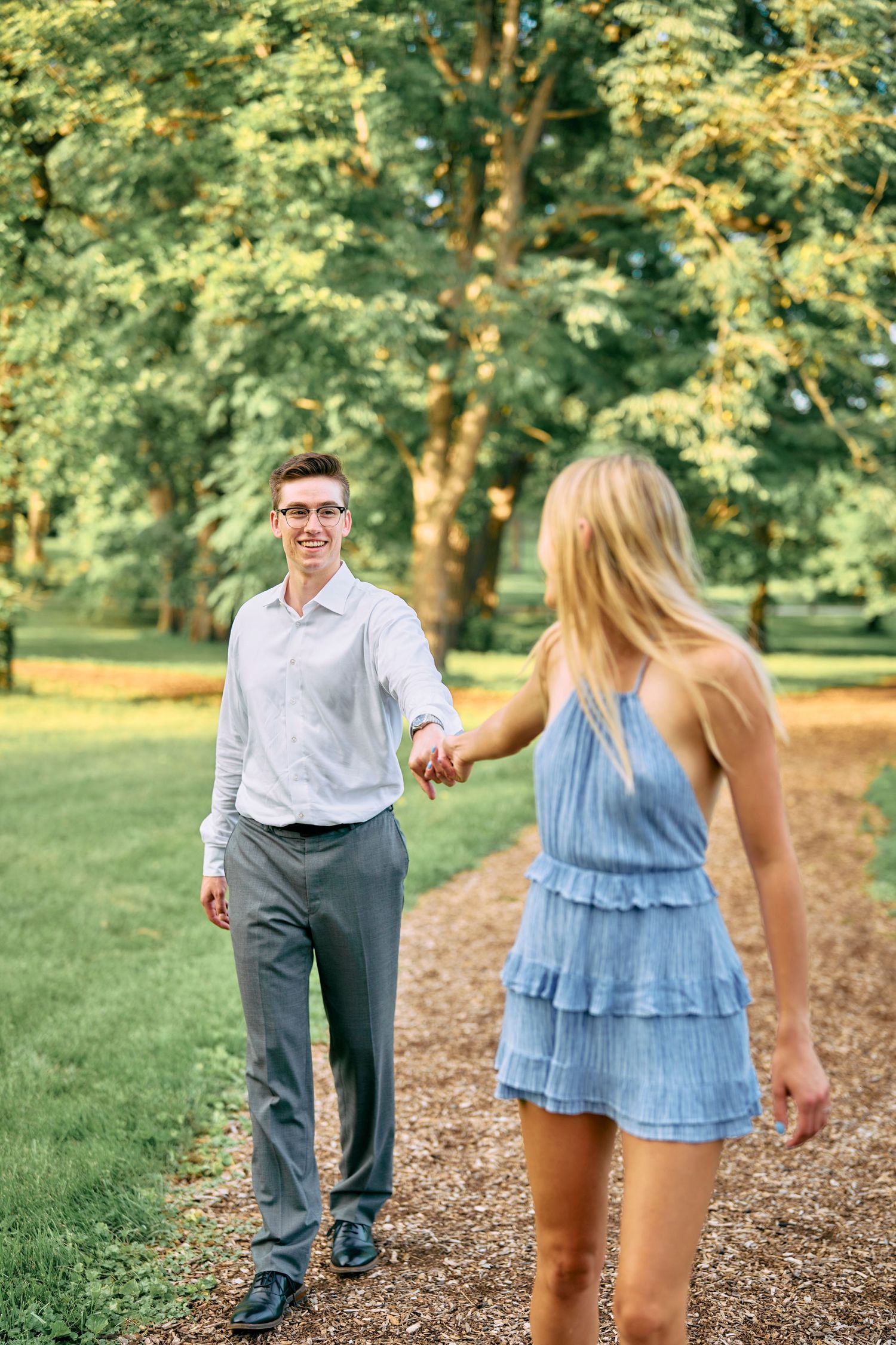 A couple walks together on a tree-lined path during a summer day, with one wearing a blue dress and the other in business casual.
