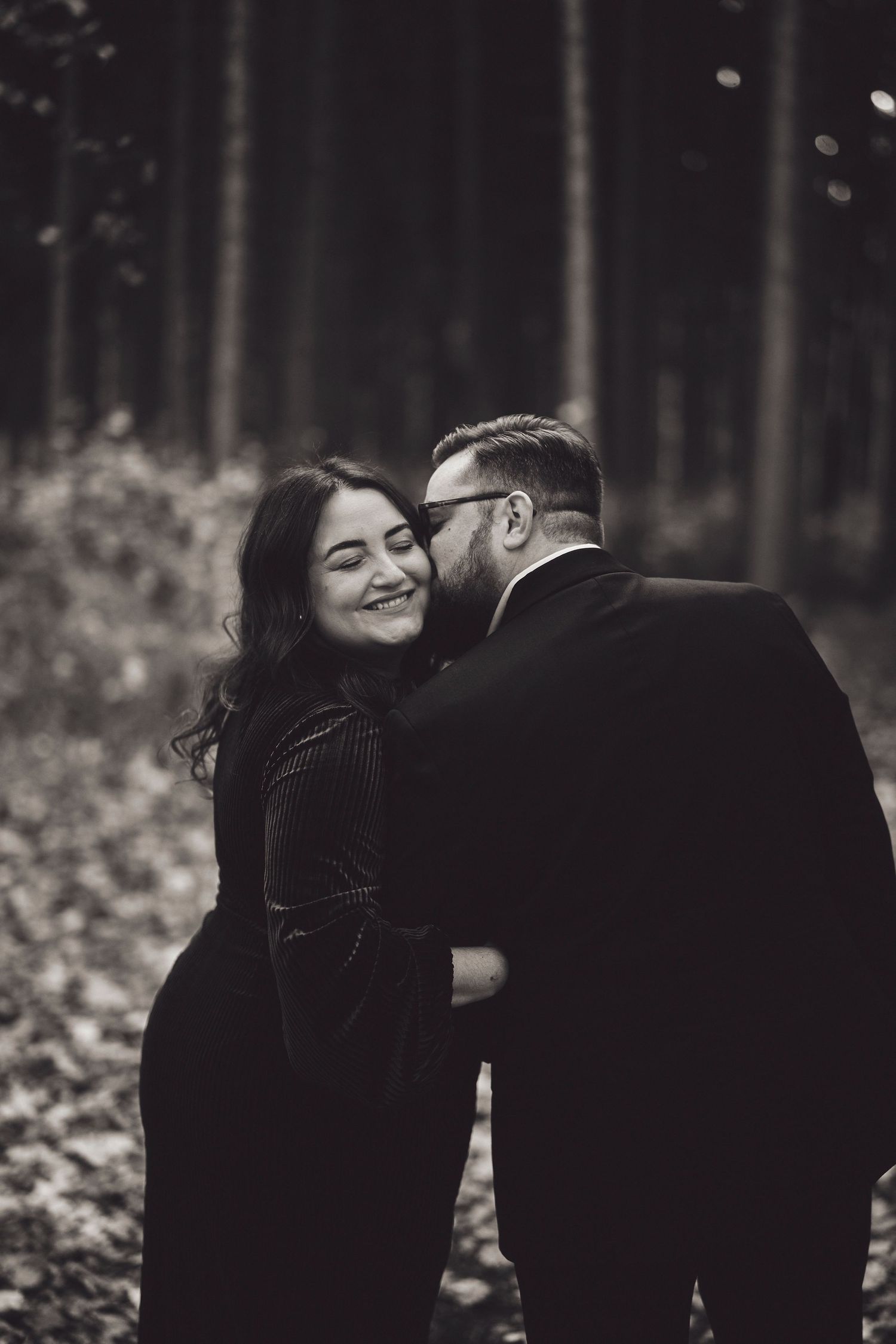 A romantic black and white photo of a couple embracing and smiling in a forest setting.