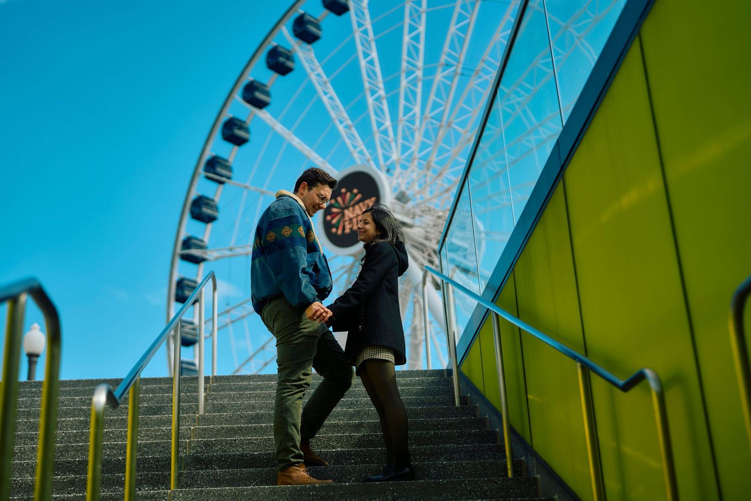A couple embraces on concrete steps with Seattle Great Wheel ferris wheel in background against blue sky.