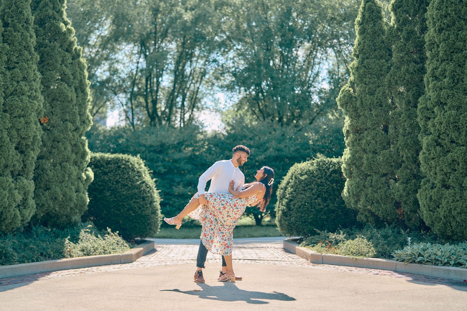 A couple shares a romantic moment on a garden path lined with tall cypress trees on a sunny afternoon.