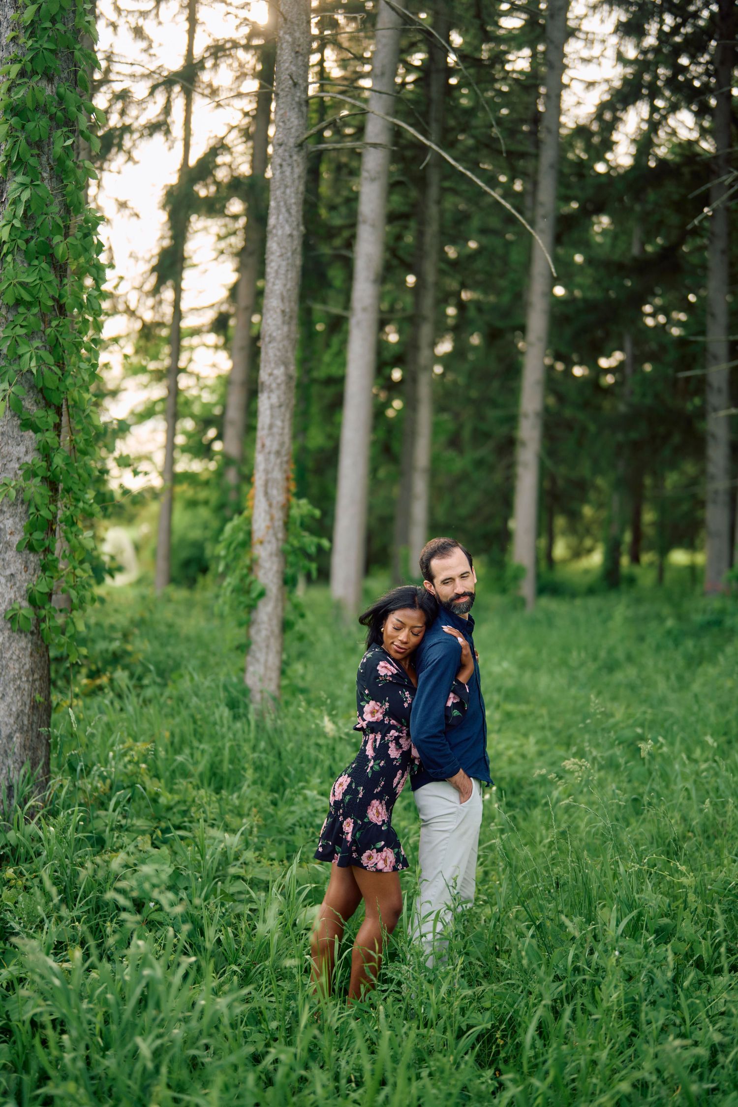 A couple stands close together among tall evergreen trees in a lush green forest during golden hour.