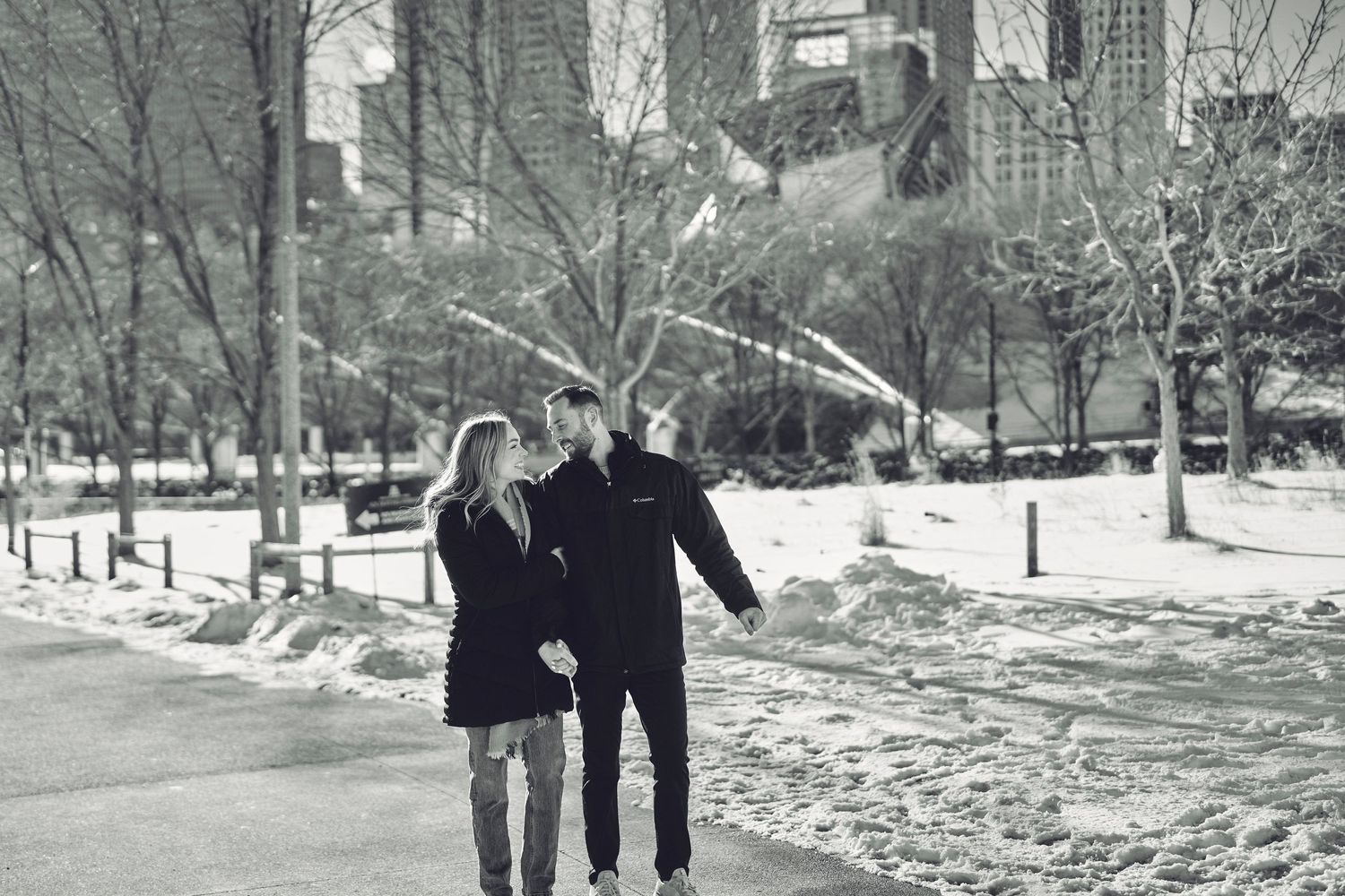A couple walking together through a snowy winter landscape in Central Park with city buildings visible in background.