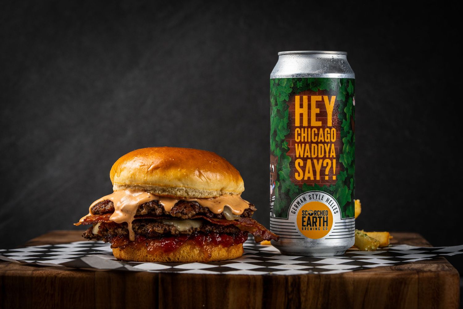 Loaded burger with melted cheese on checkered surface beside craft beer can.