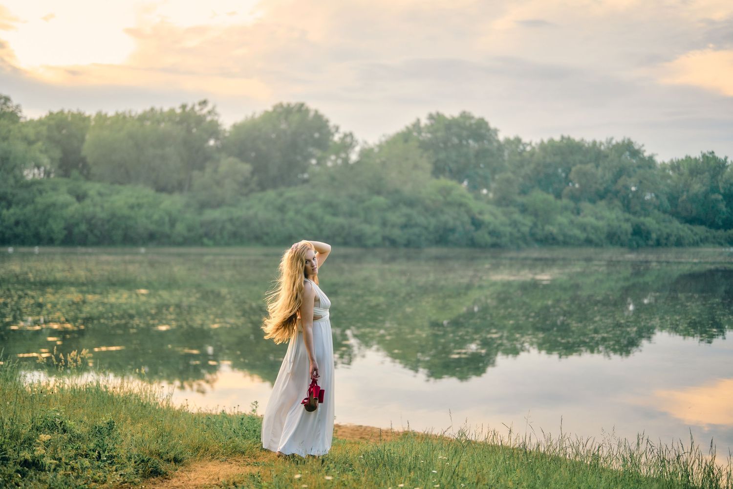 Person in a white dress stands by a tranquil lake surrounded by trees at sunset with reflections in the calm water.
