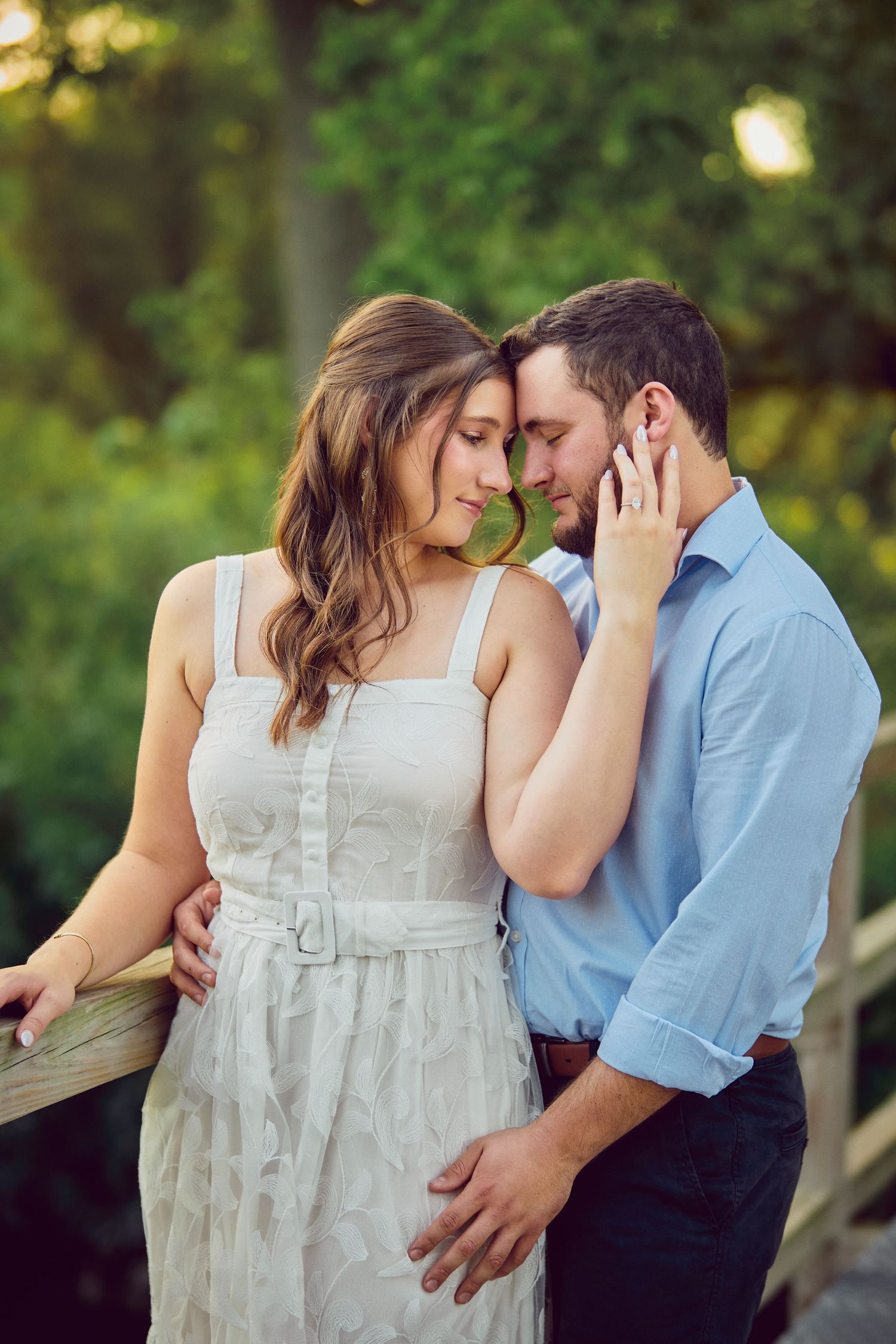 A romantic couple shares an intimate moment outdoors, she in a white dress and he in a blue shirt.