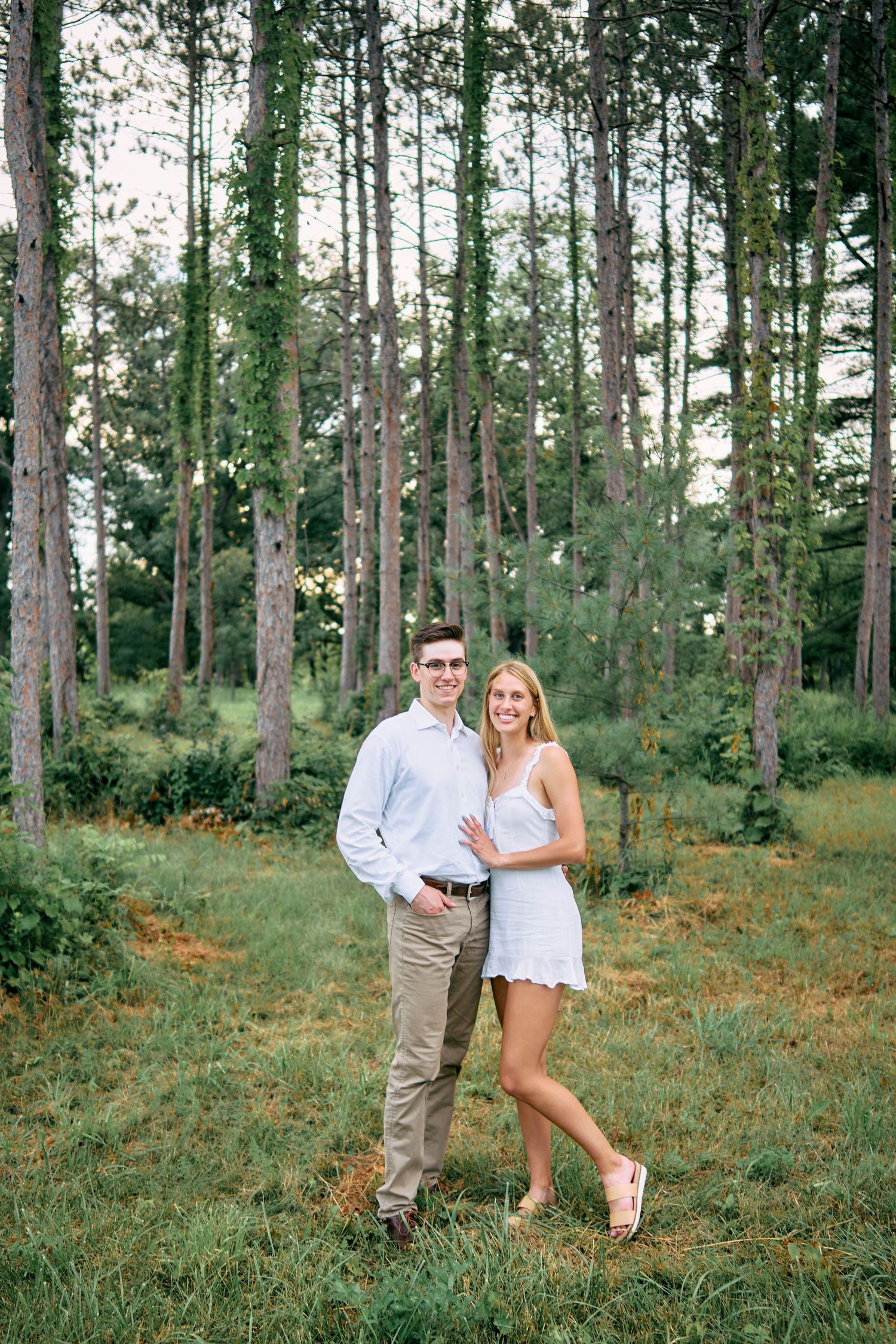 A couple poses together in a sunlit forest wearing coordinated light-colored summer outfits.