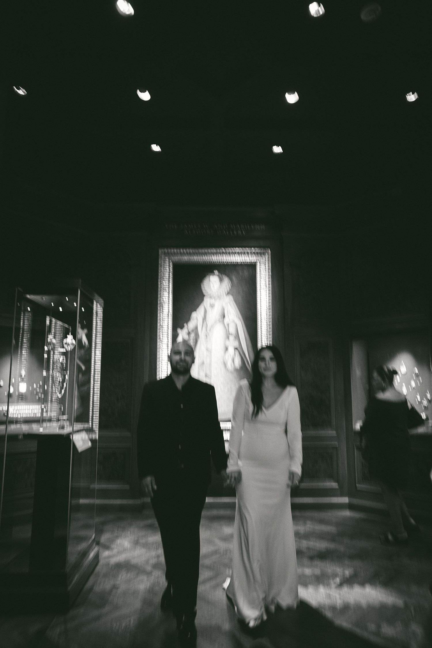 Black and white photo showing a couple walking together in a formal setting with artwork and ornate frames on the walls.