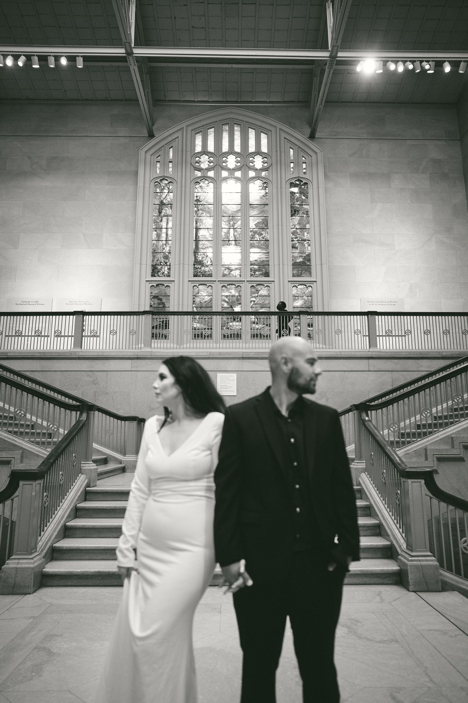 Black and white wedding photo inside architectural building with grand staircase and stained glass window in background.