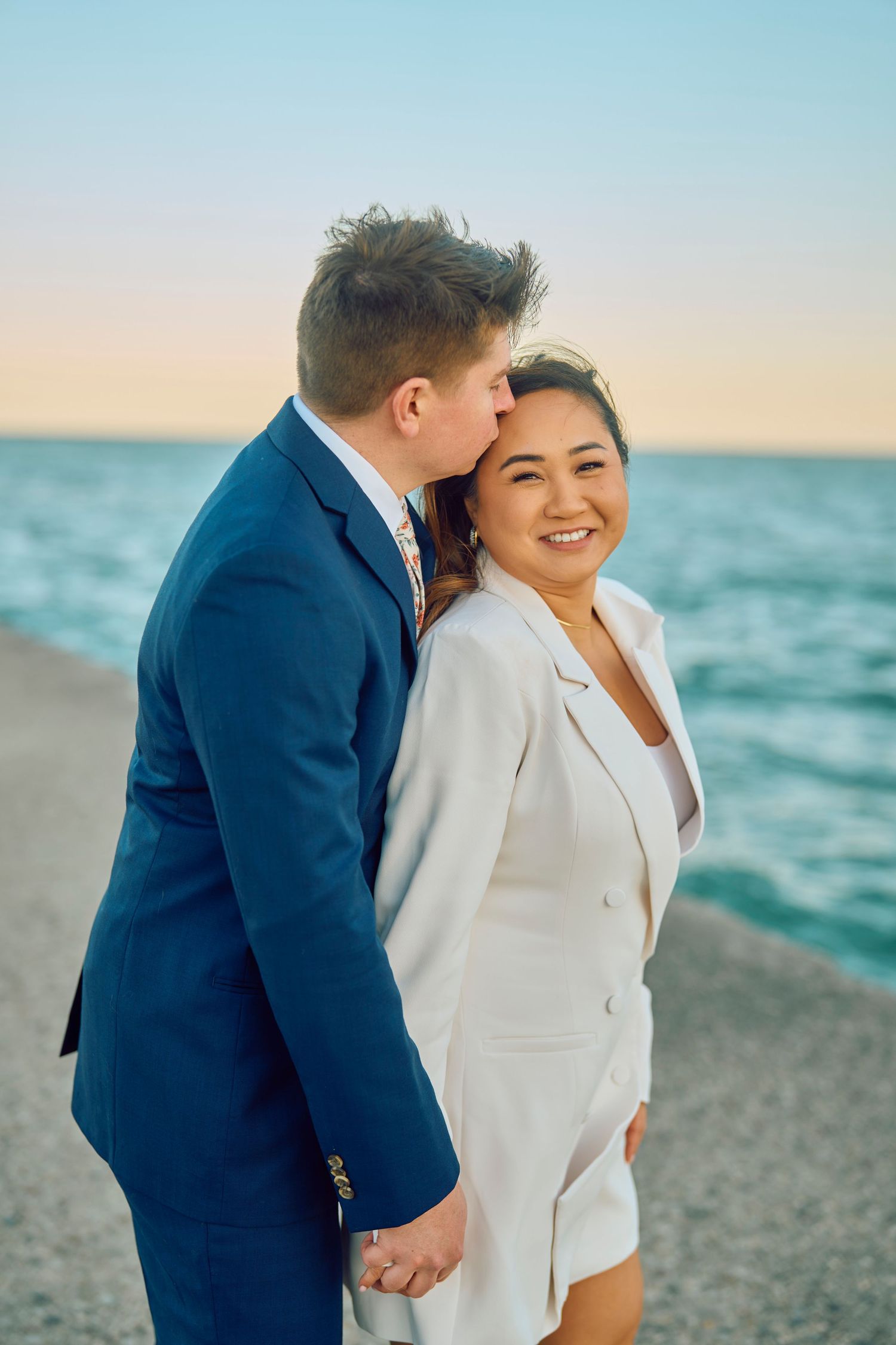 A romantic couple in formal attire share a tender moment on a beach at sunset, with blue ocean waters in the background.