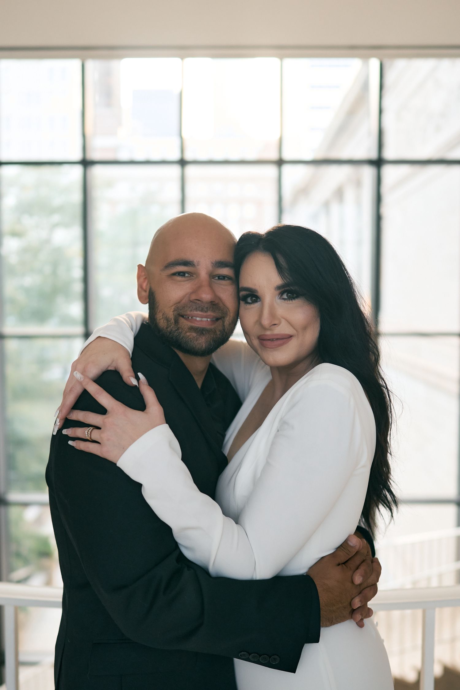 A couple embraces in front of a sunlit window wearing matching black and white formal attire.