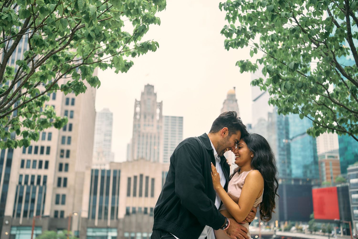A romantic couple embraces in front of a cityscape with tall buildings and green trees framing the urban scene.
