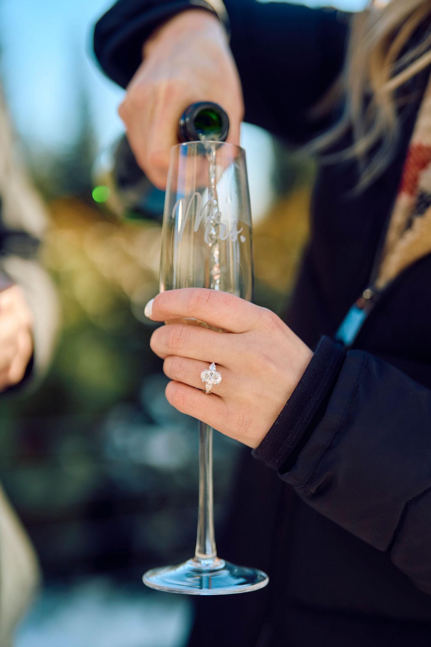 Someone wearing black pours champagne into an elegant flute glass while showing off a diamond engagement ring.