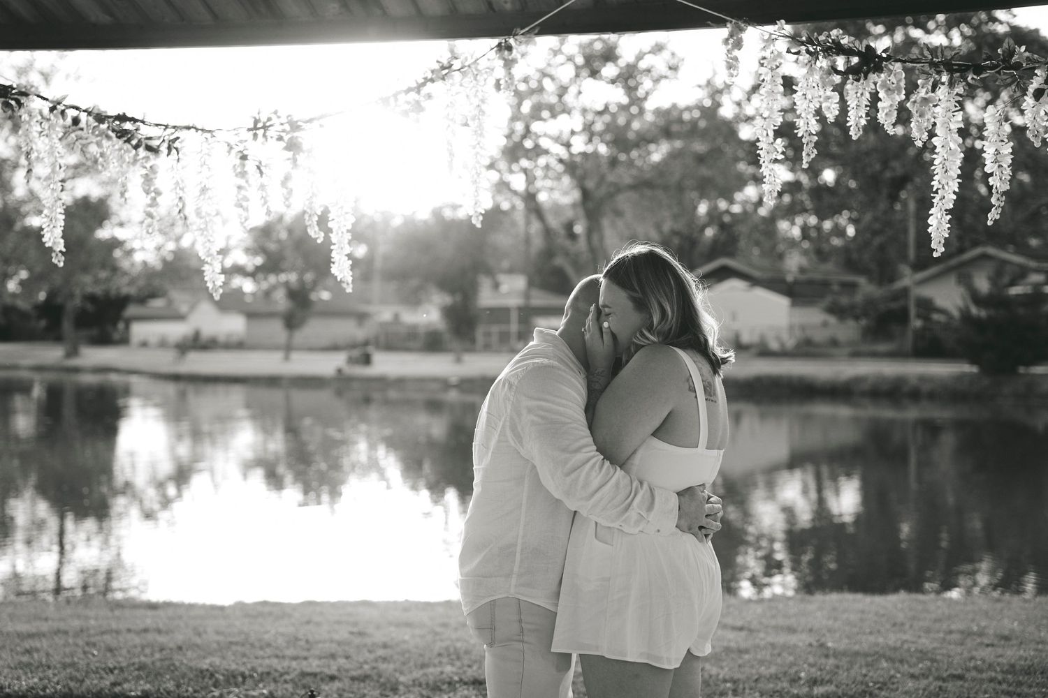 Black and white photo of a romantic couple embracing by a lake at sunset with trees and reflections on the water.