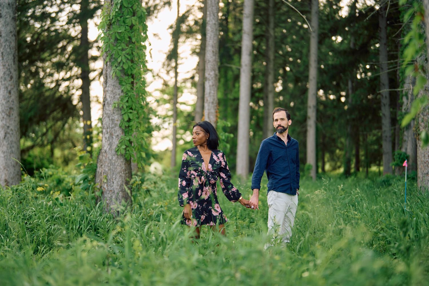 A couple walks hand in hand through a lush green forest setting with ivy-covered trees.