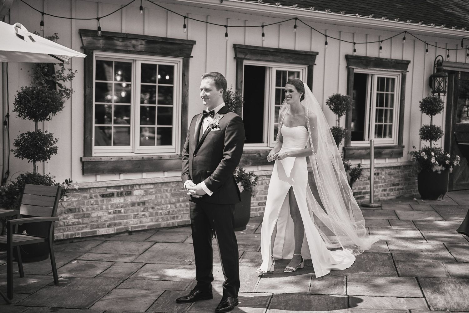 A couple in formal wedding attire share their first look moment on a stone patio outside a rustic venue.