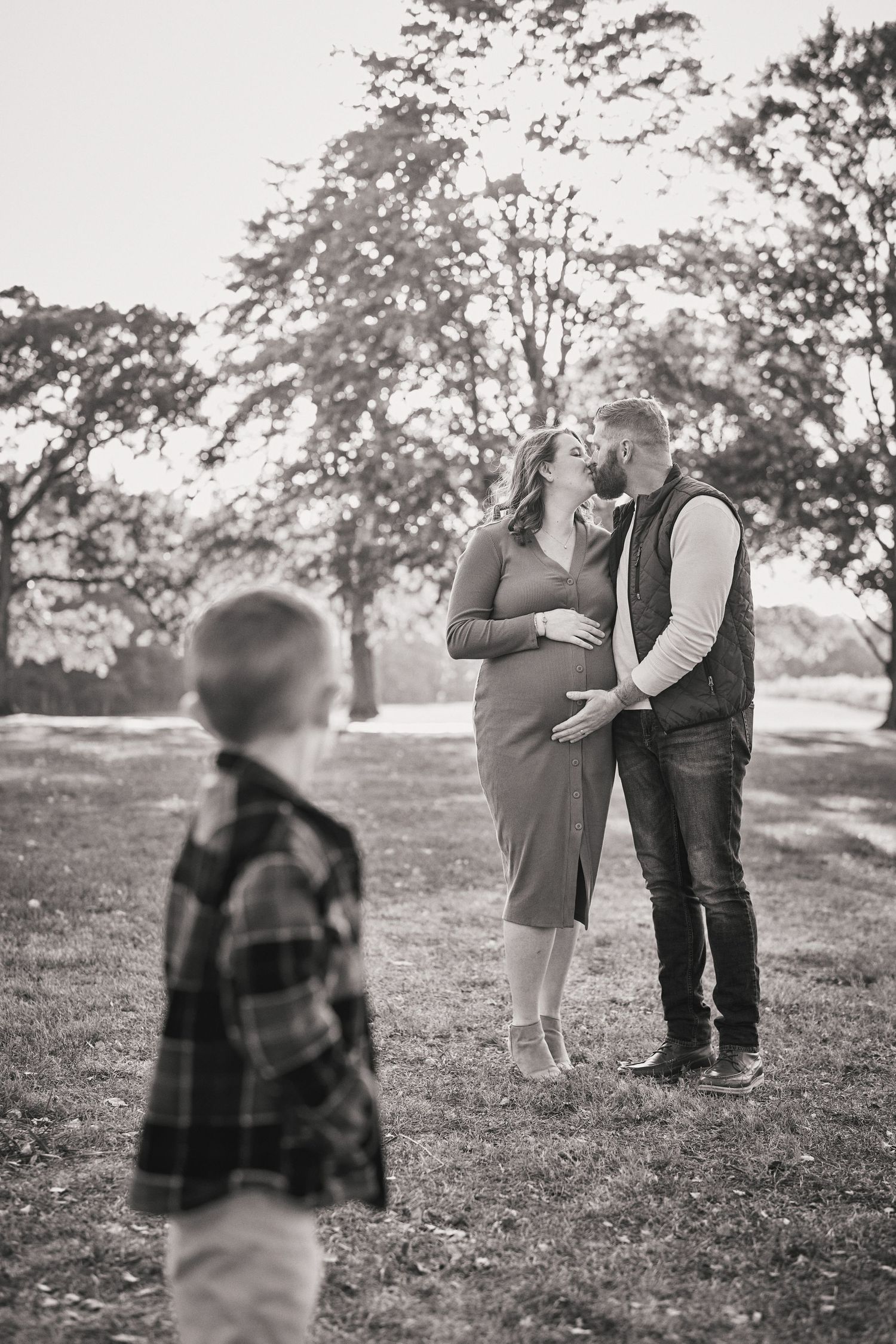 Black and white photo of couple embracing outdoors while young child in plaid shirt watches from a distance.
