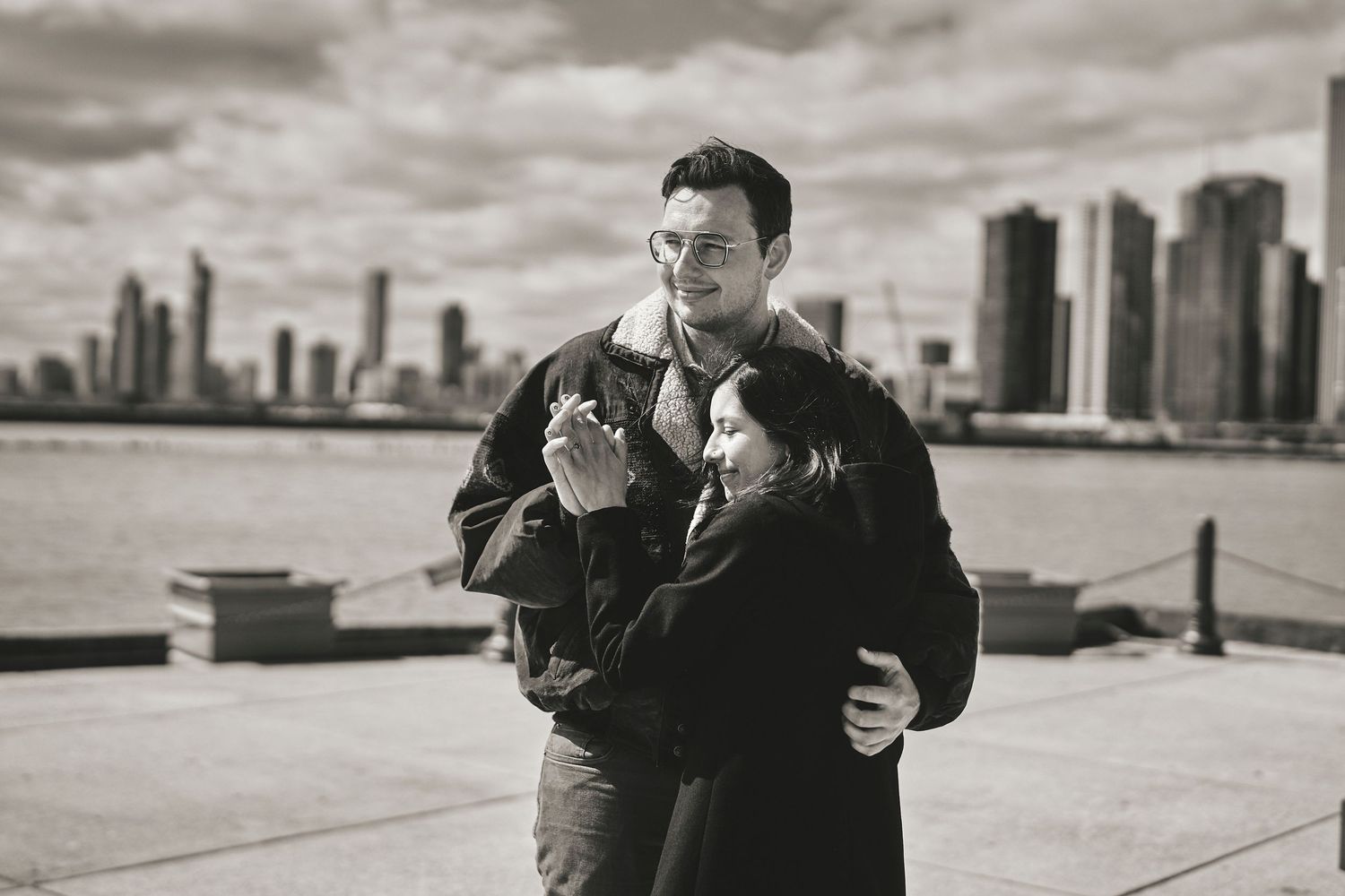 Black and white photo of two people embracing on a waterfront promenade with city skyline in background.