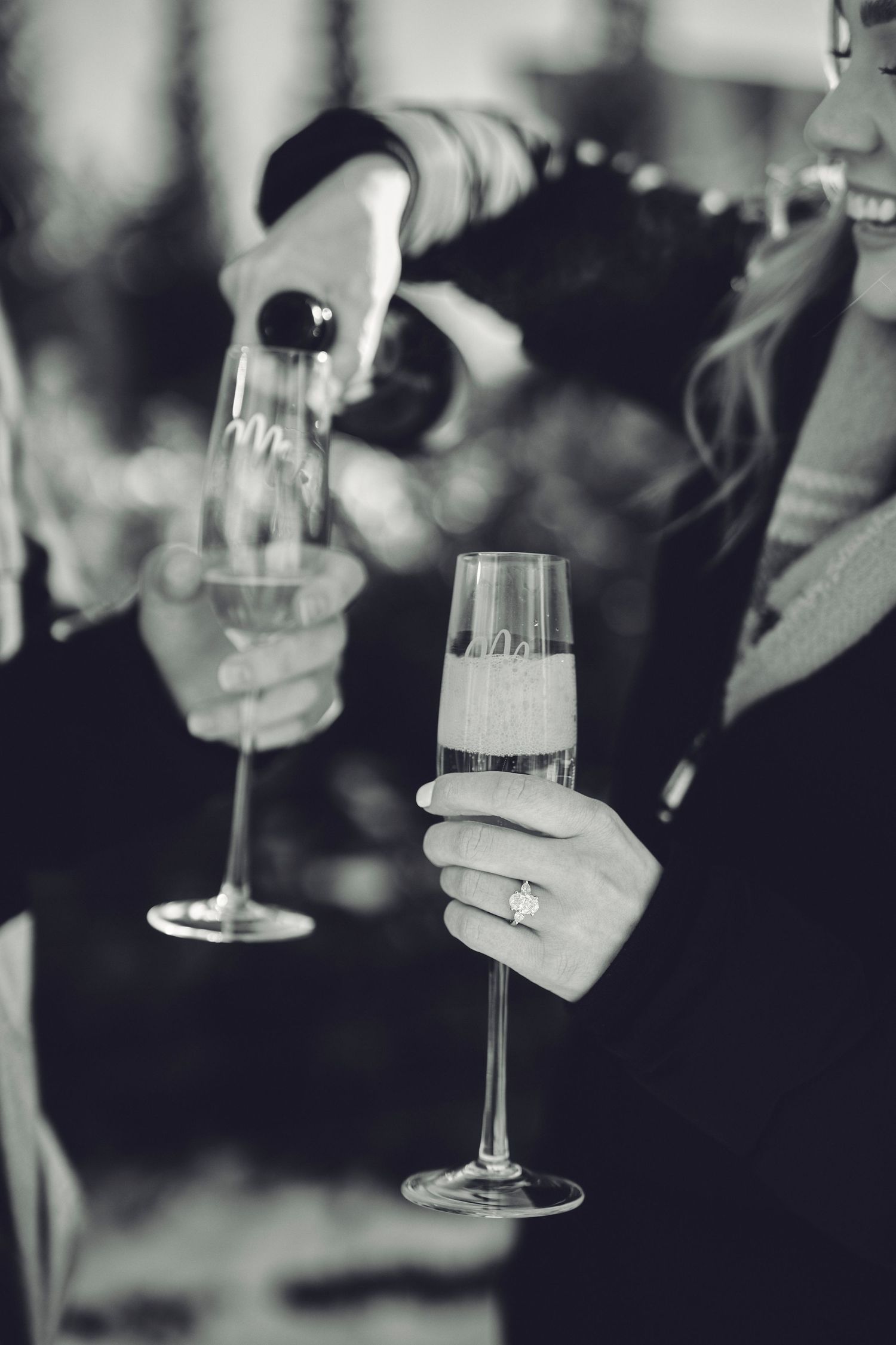 A black and white photo of champagne being poured between two elegant flute glasses held by hands wearing rings.
