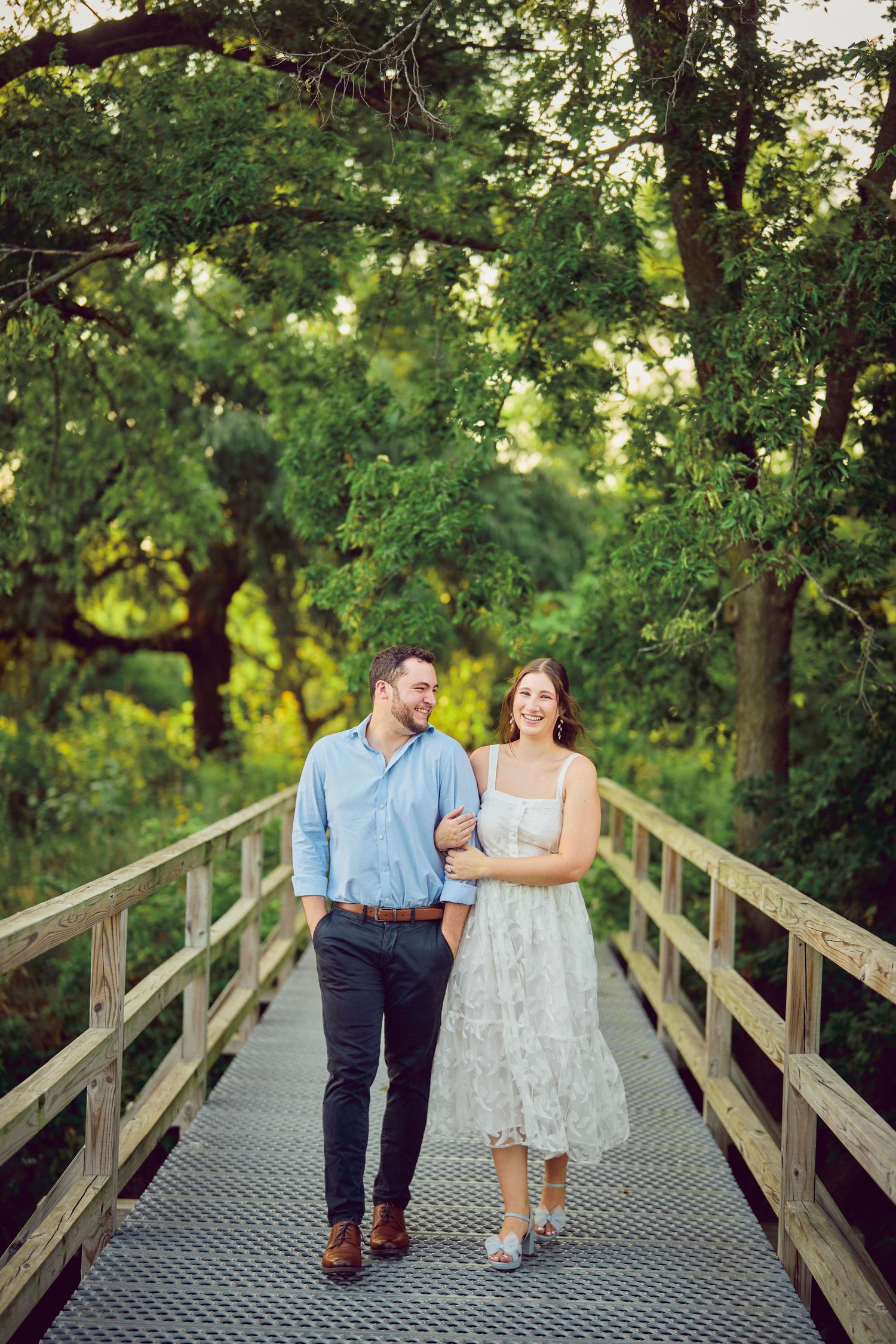 A couple walks together on a wooden footbridge surrounded by lush green trees in a natural park setting.