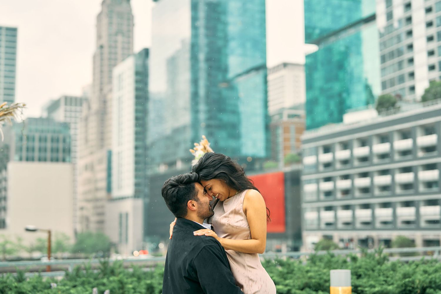A romantic couple shares a tender embrace against the backdrop of modern city skyscrapers in an urban garden setting.