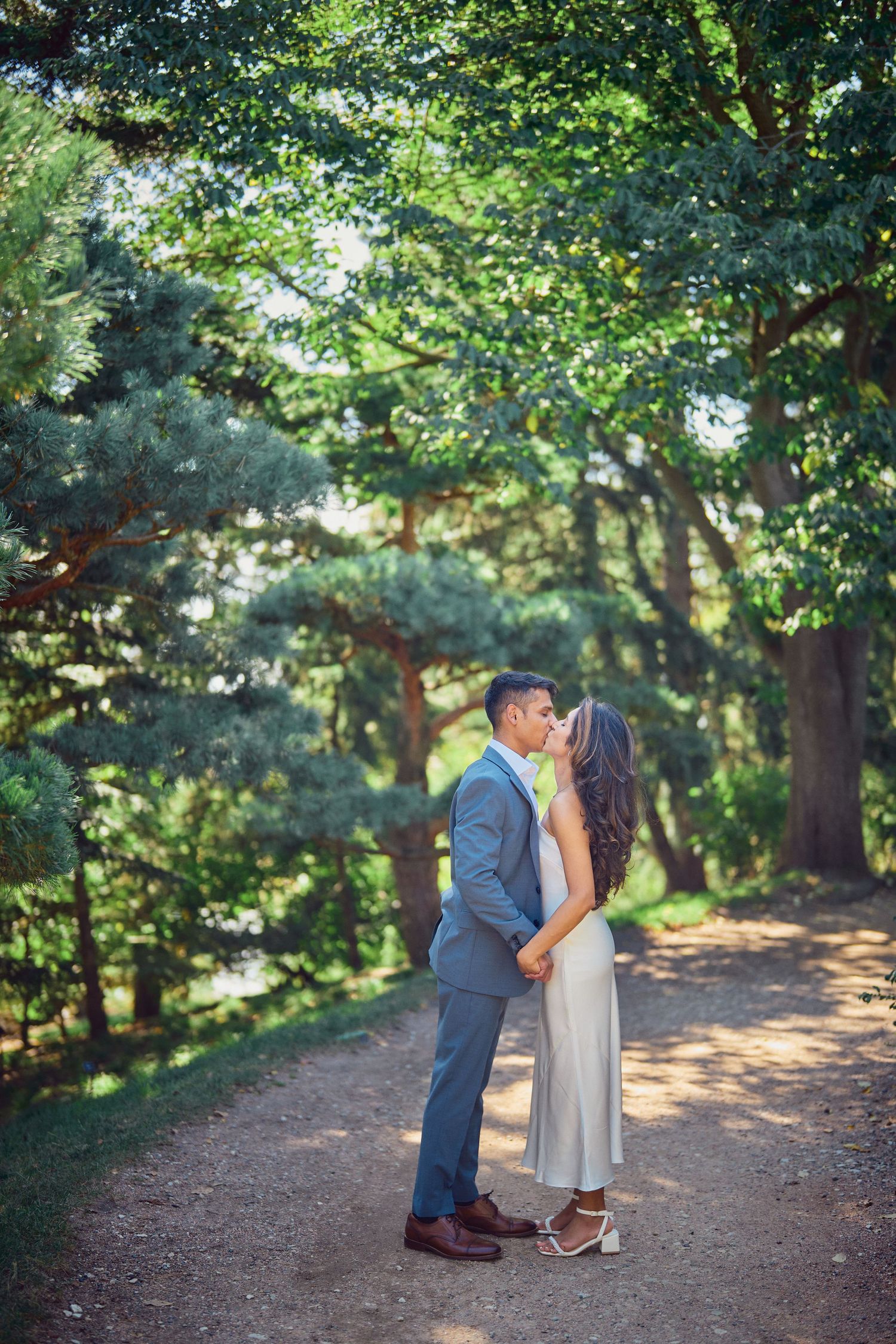 A couple shares a romantic moment on a sunlit forest path surrounded by lush green trees and dappled light.