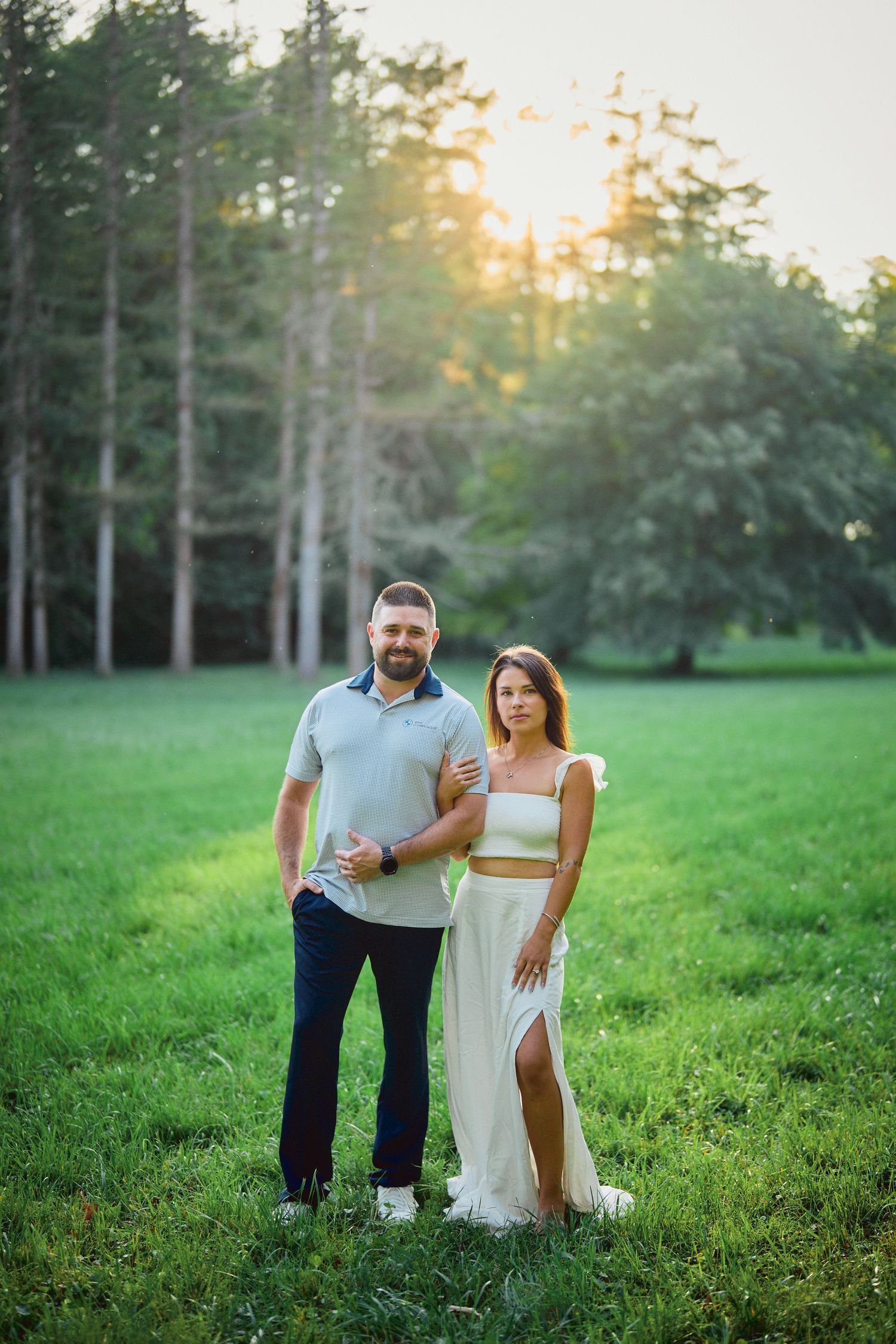 A couple stands together in a grassy field at sunset with tall pine trees in the background.