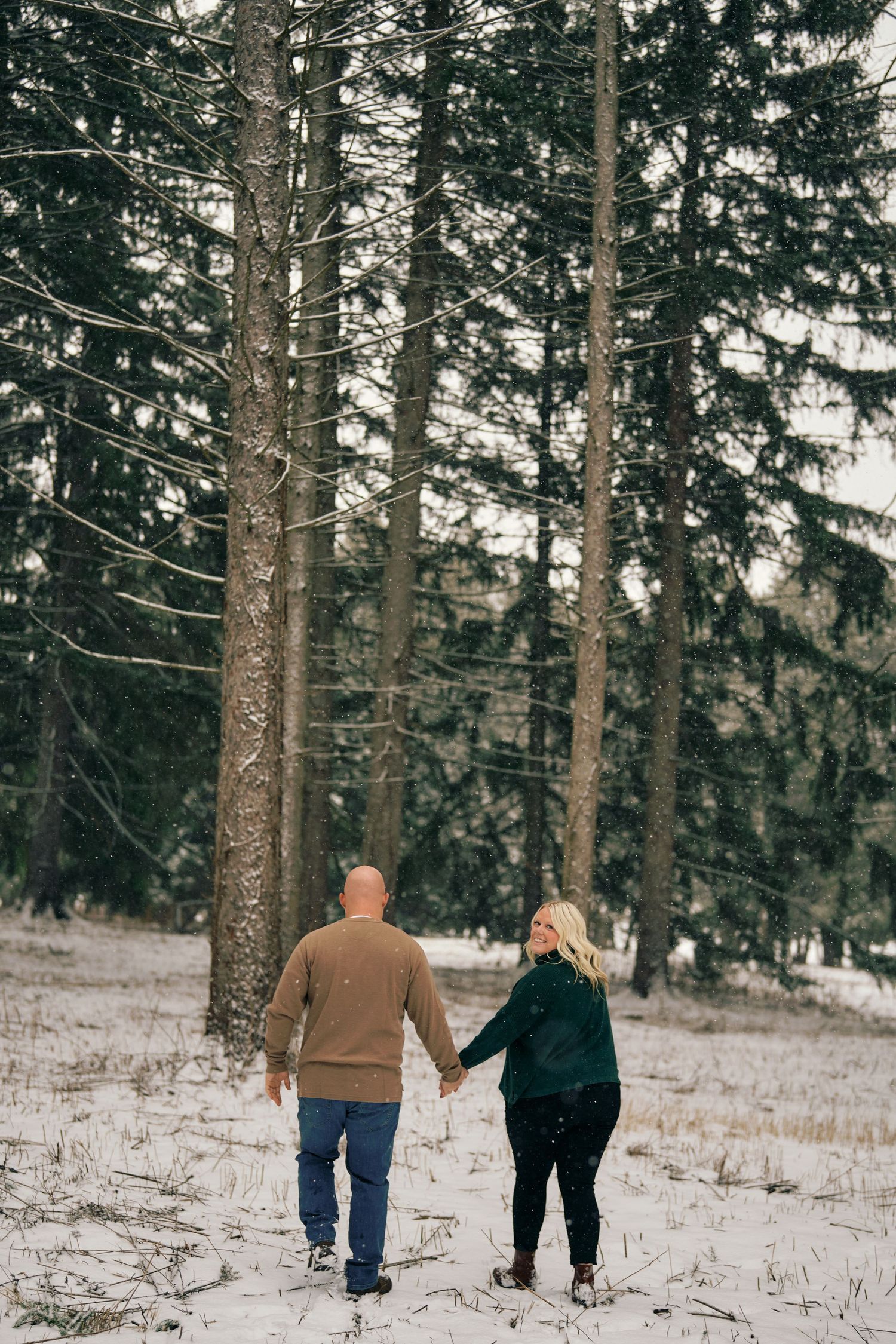 A couple walking hand in hand through a snowy forest path surrounded by tall evergreen trees.