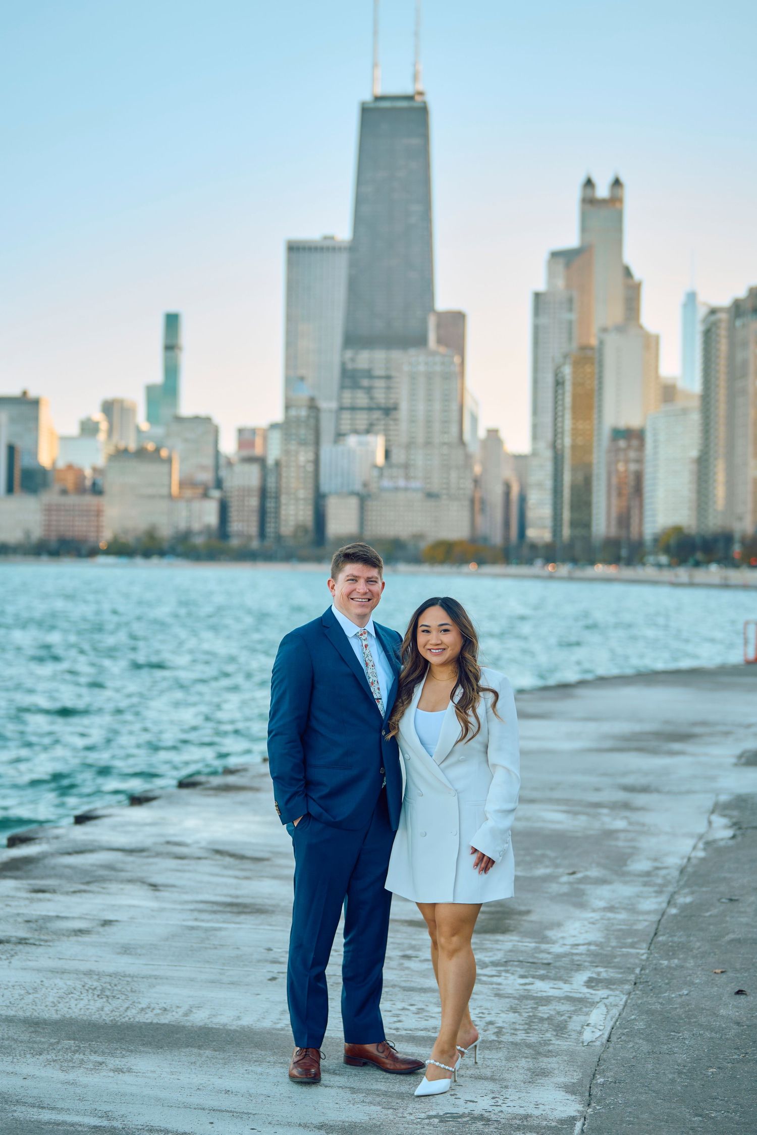 A couple poses together along Lake Michigan with the Chicago skyline featuring the iconic John Hancock building in the background.