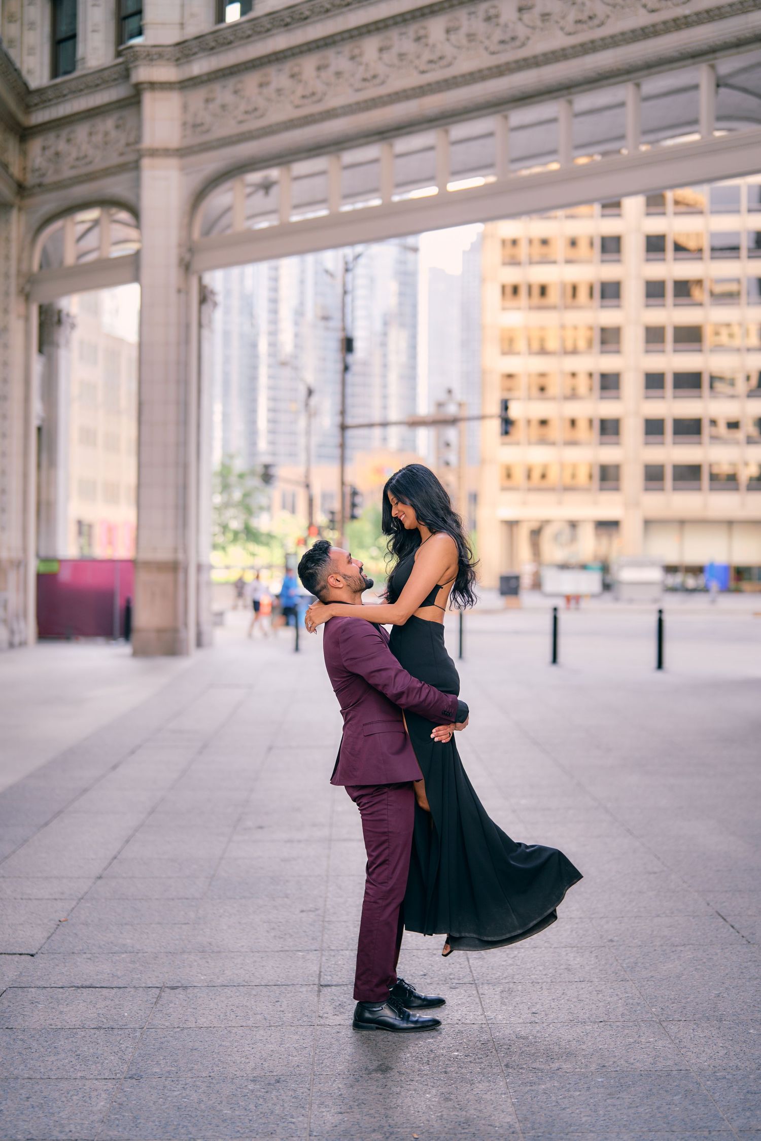 A couple shares a romantic moment in front of an elegant urban building, with the man lifting his partner in a burgundy suit.