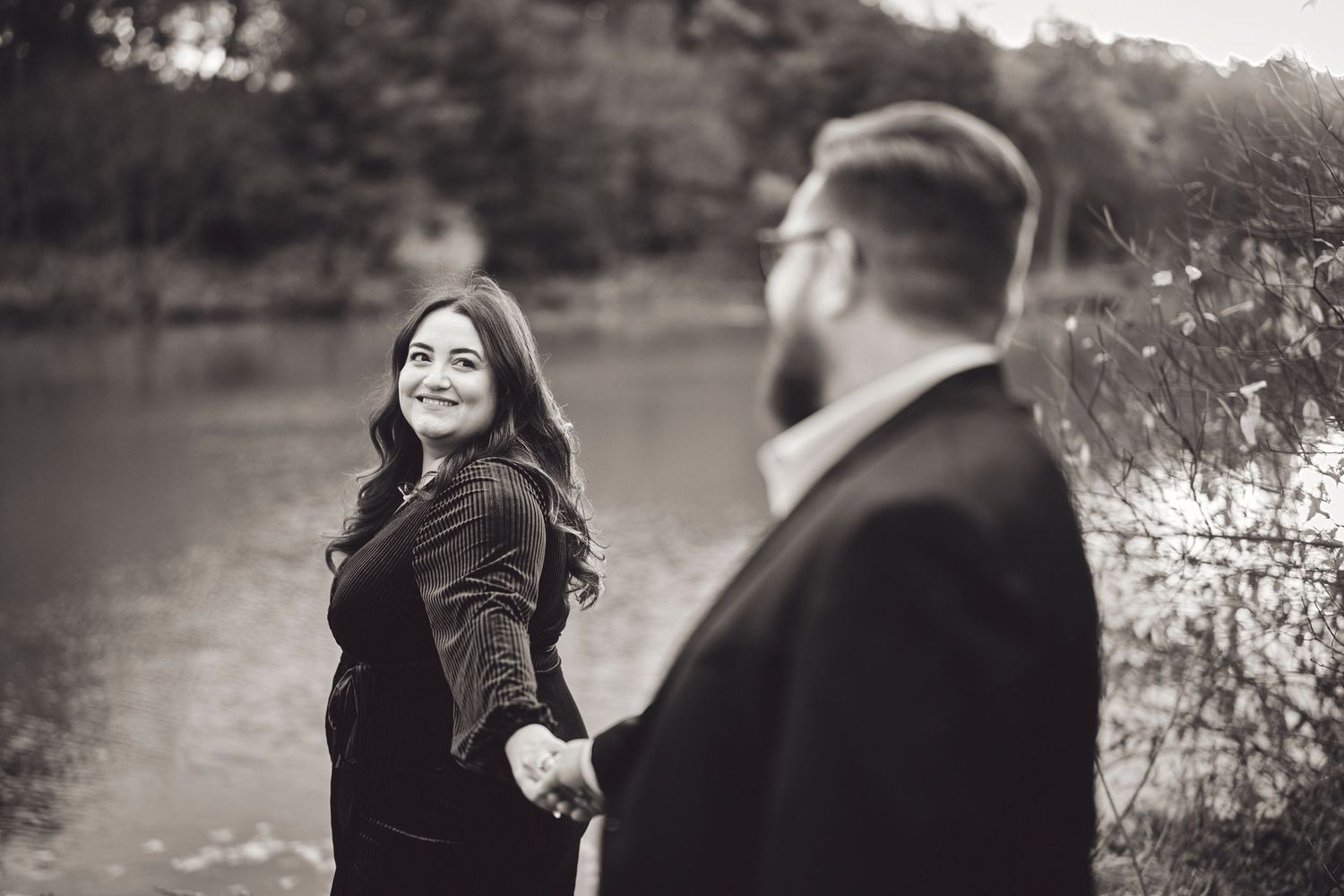 A couple shares a romantic moment by a lake in black and white, with one leading the other along the water's edge.