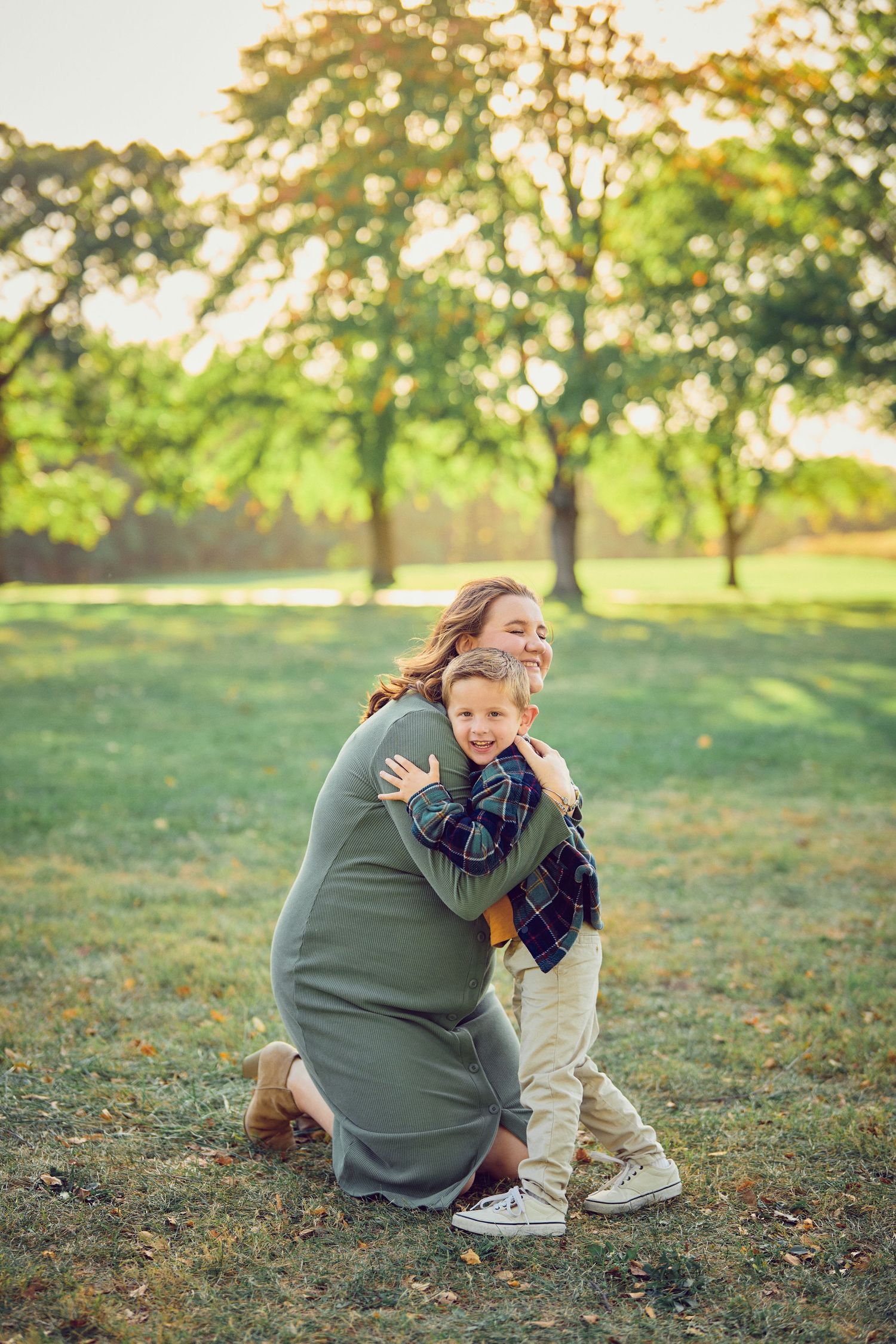A tender embrace shared in a sunlit park with green trees and golden light filtering through the leaves at sunset.