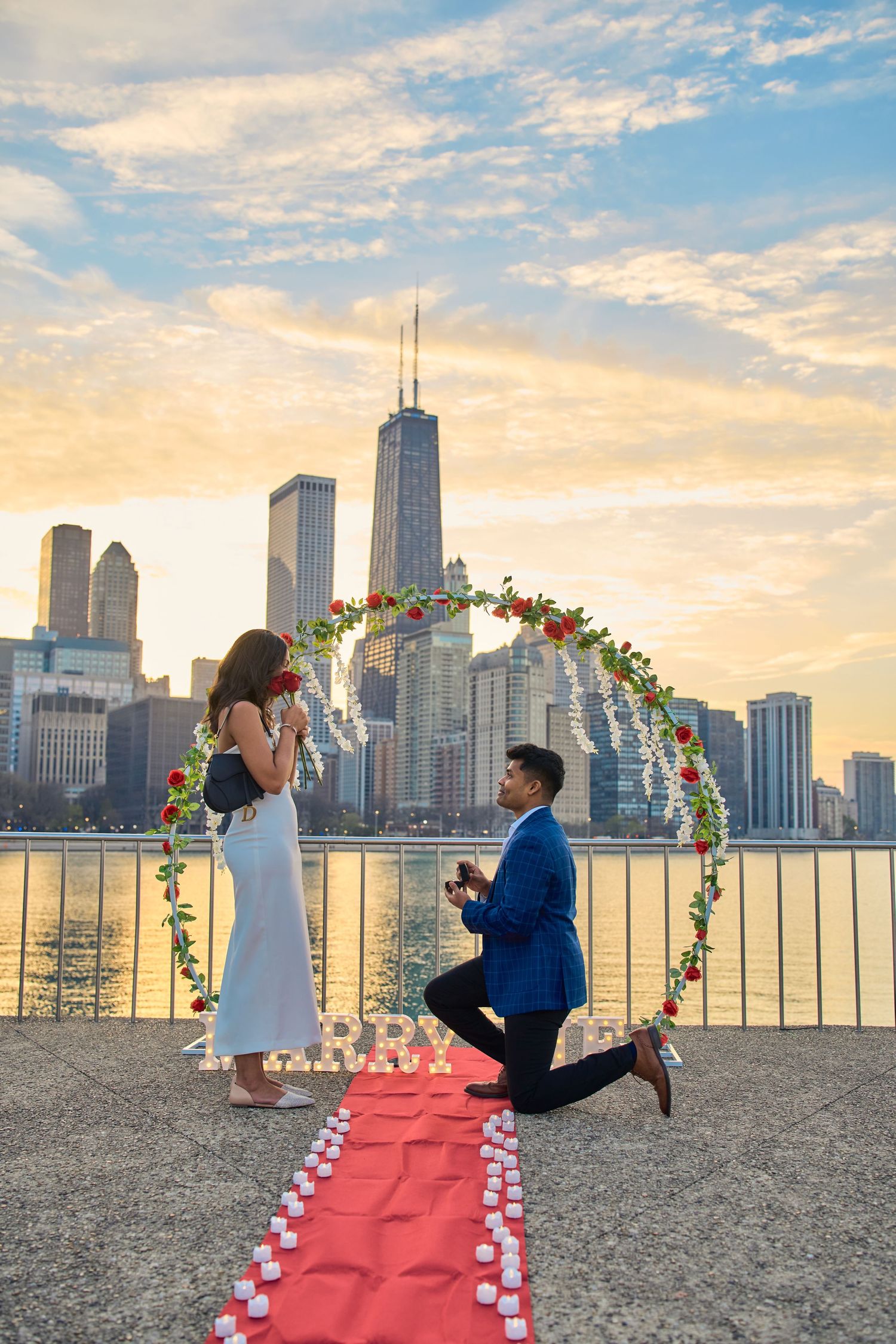 Marriage proposal at sunset with Chicago skyline backdrop, floral arch and red carpet lined with candles.