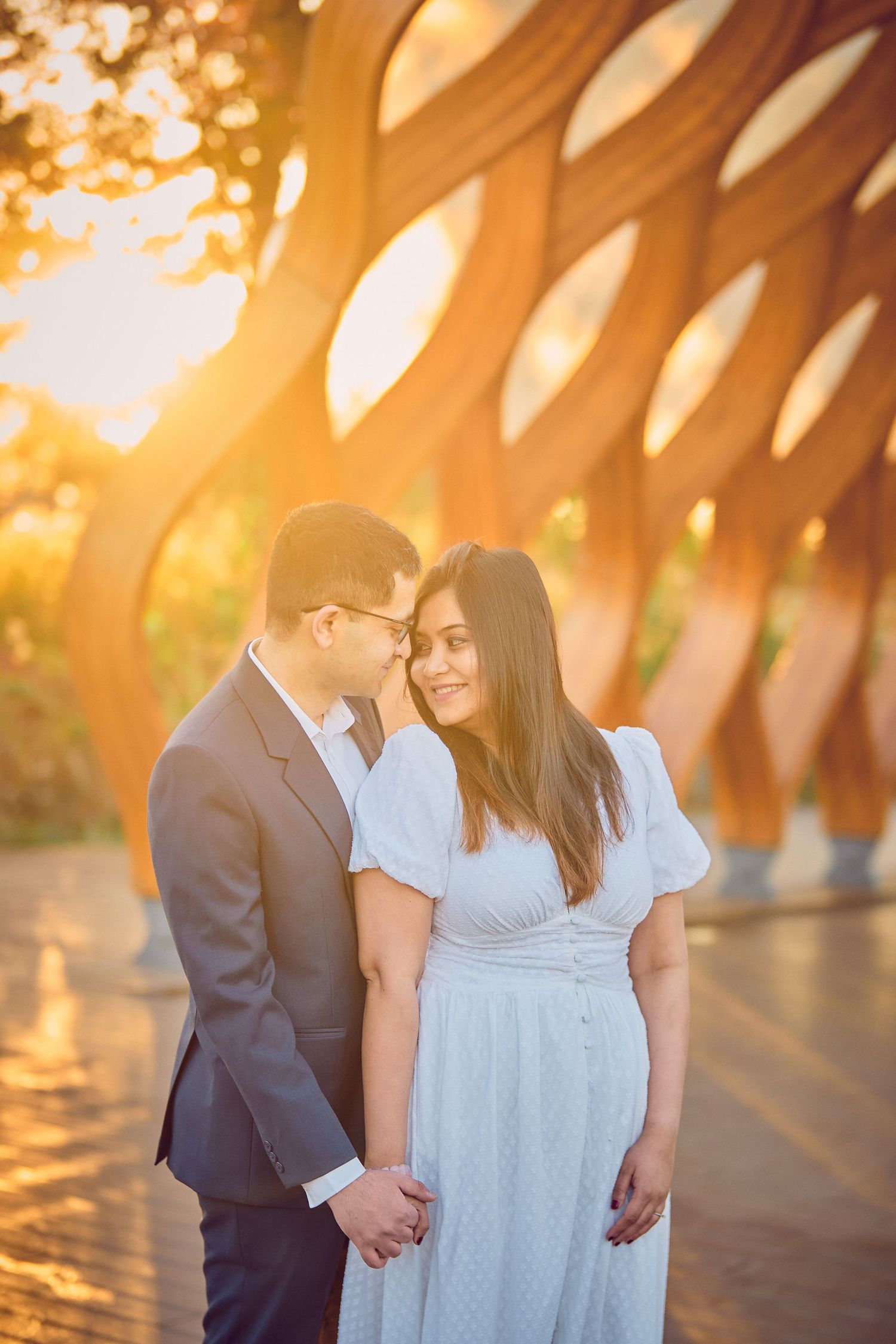 A romantic sunset engagement photo with a geometric architectural backdrop creating dramatic golden lighting patterns.