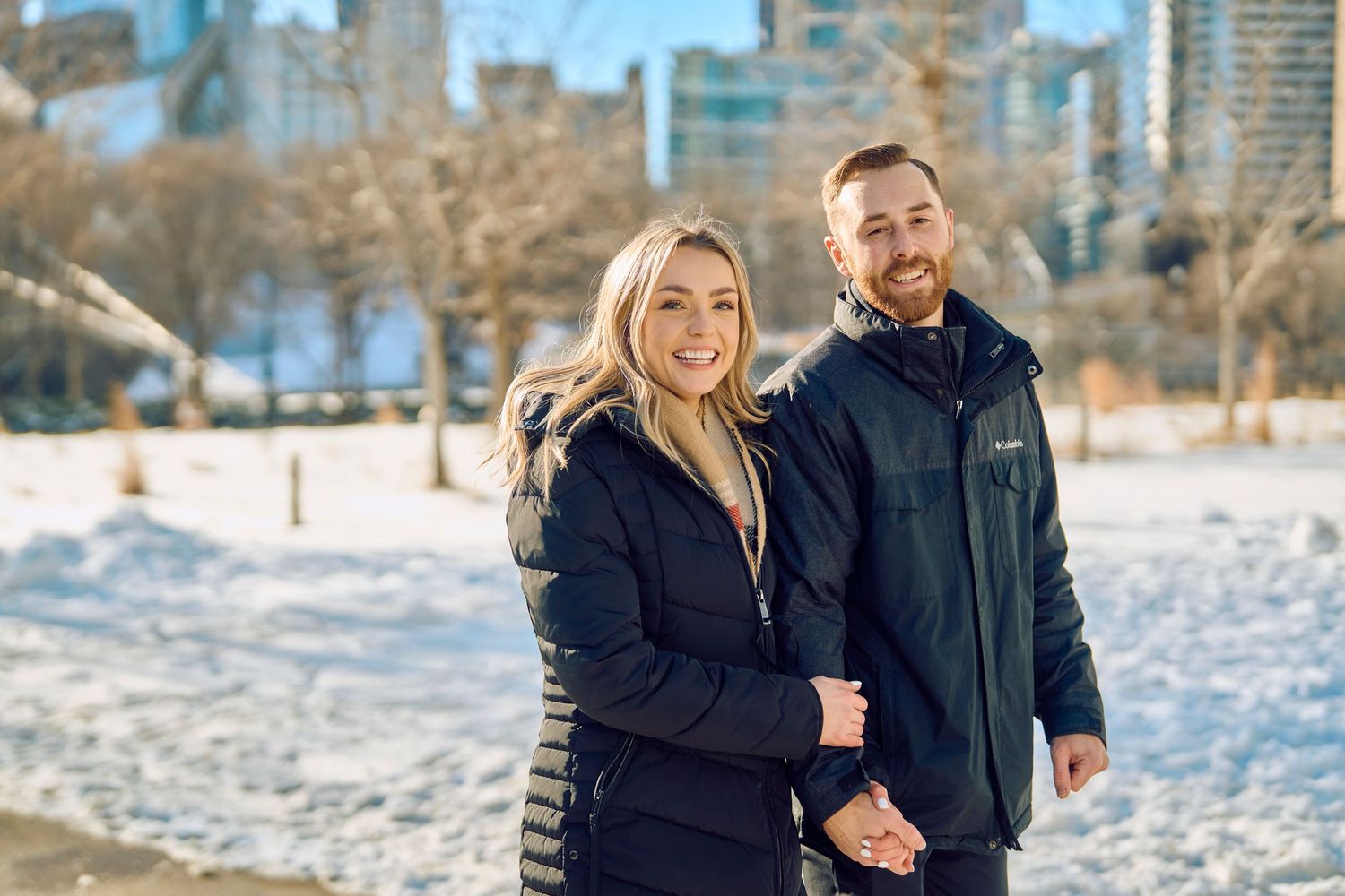 Two people in winter coats pose together on a snowy day with city buildings visible in the background.